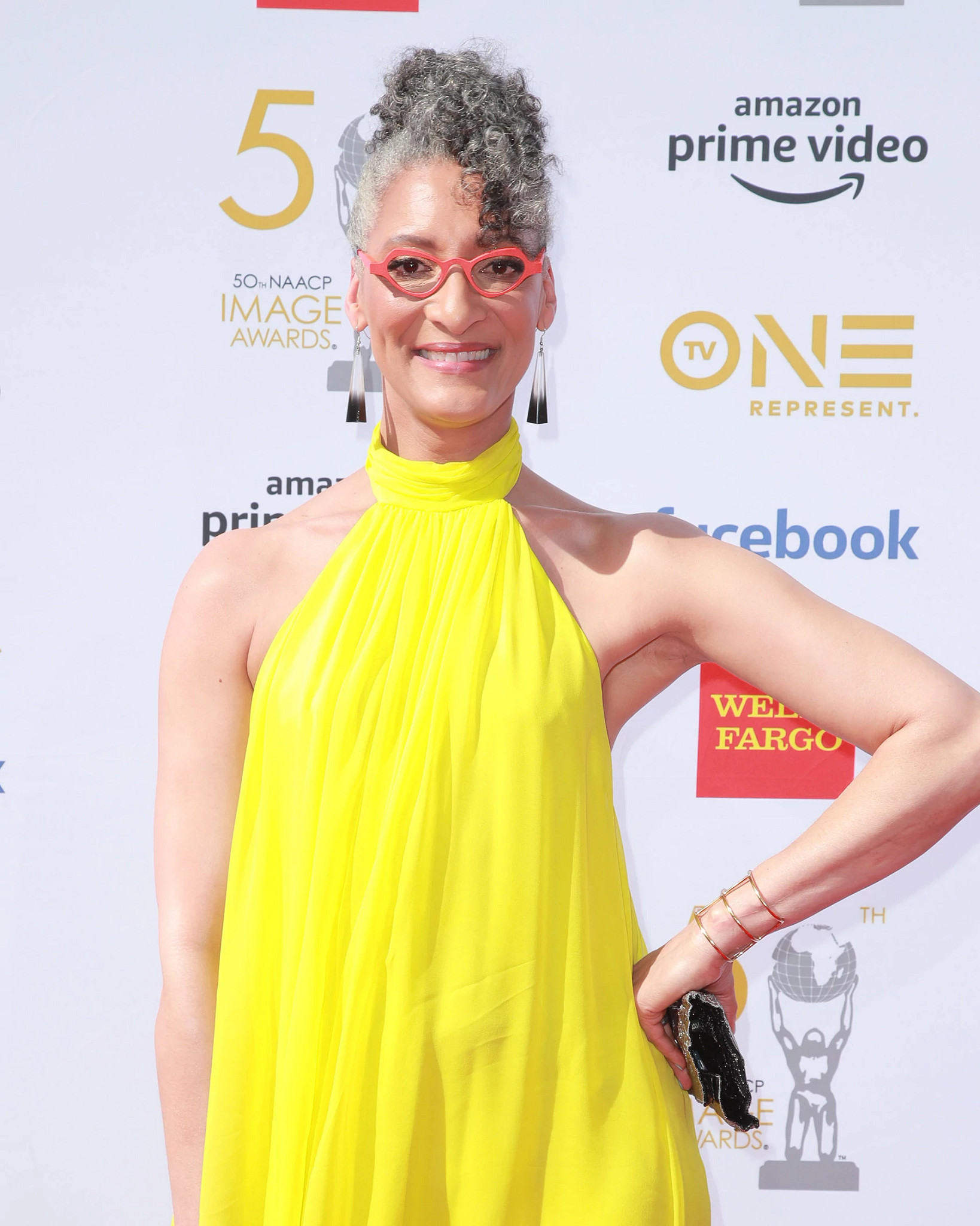 Carla Hall wearing a bright yellow sleeveless outfit and colorful eyeglasses at a public event backdrop