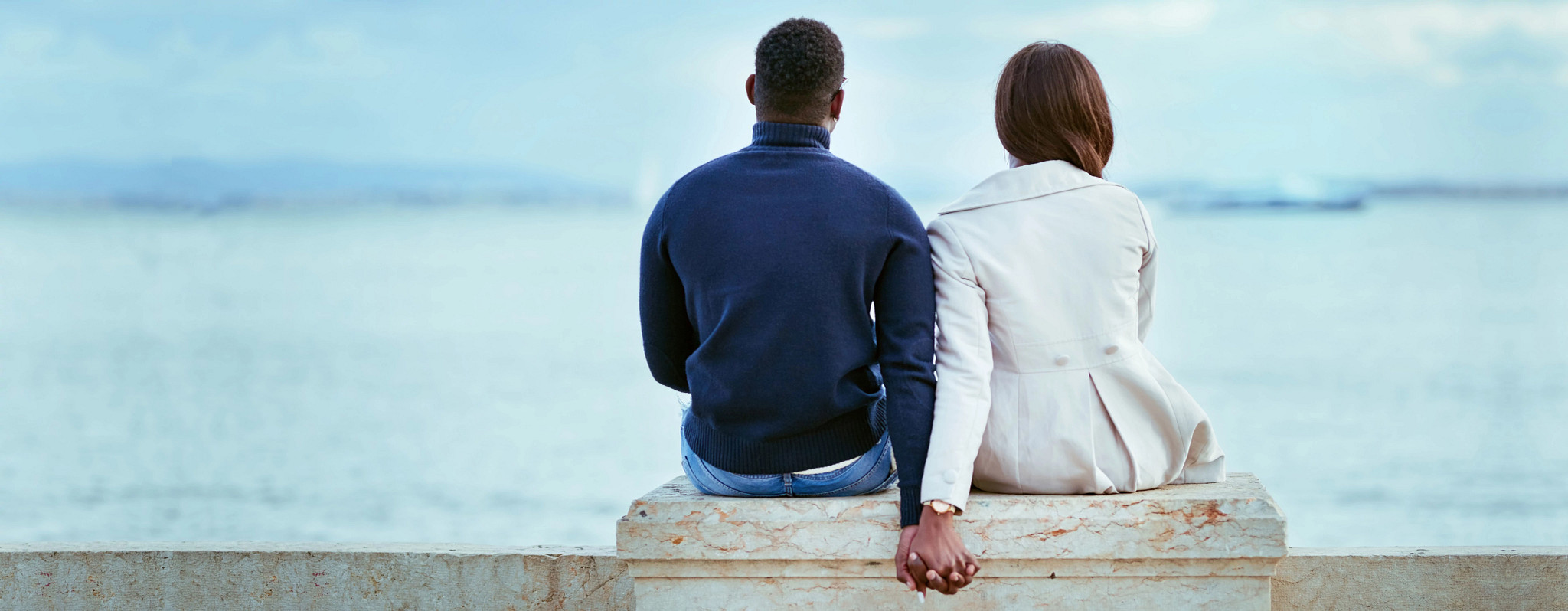 Man and woman sitting on a cement bench looking out over the ocean, holding hands