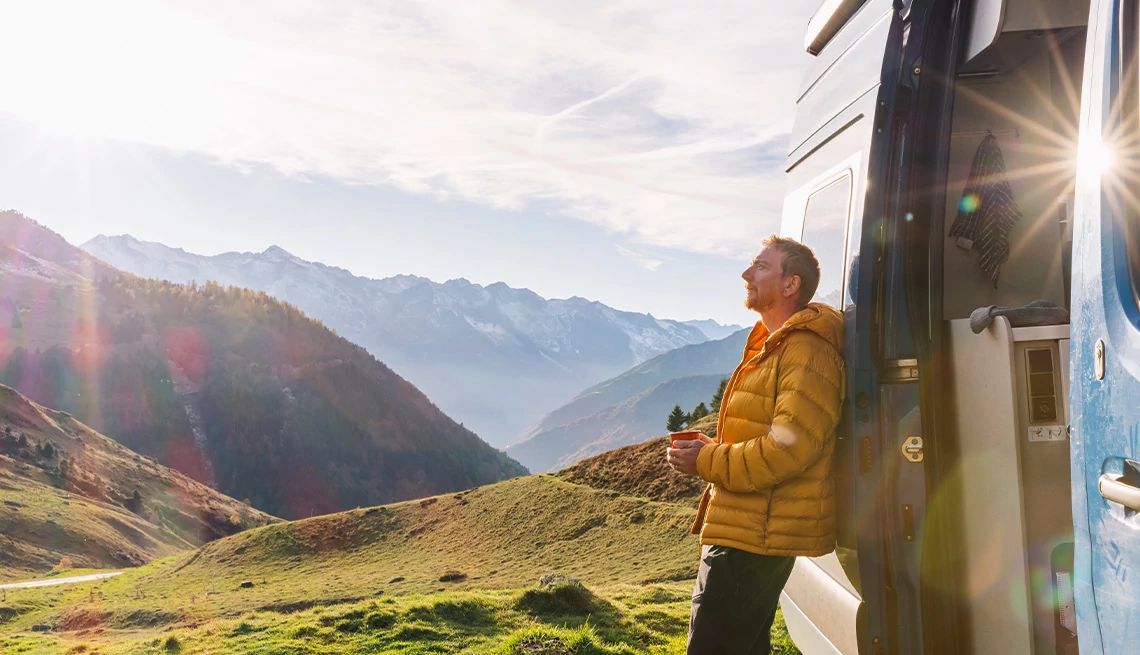 Serene looking man in yellow jacket leaning against his camping van on with a beautiful mountain view in the background