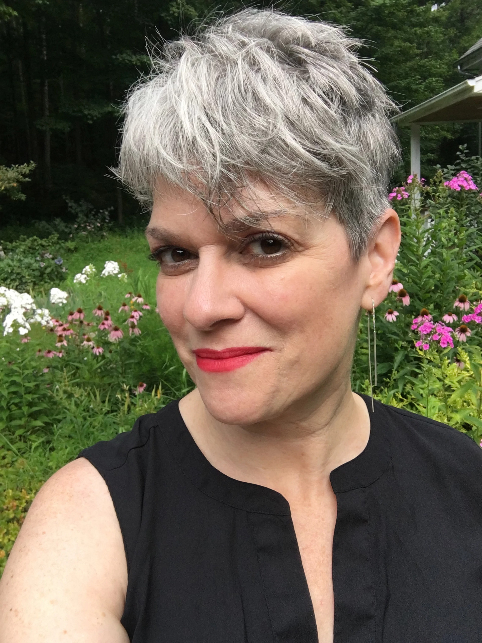 Close-up photo of Kristan Higgins with short gray hair wearing a sleeveless dark top and long earring, standing in a lush garden with blooming pink and white flowers and greenery