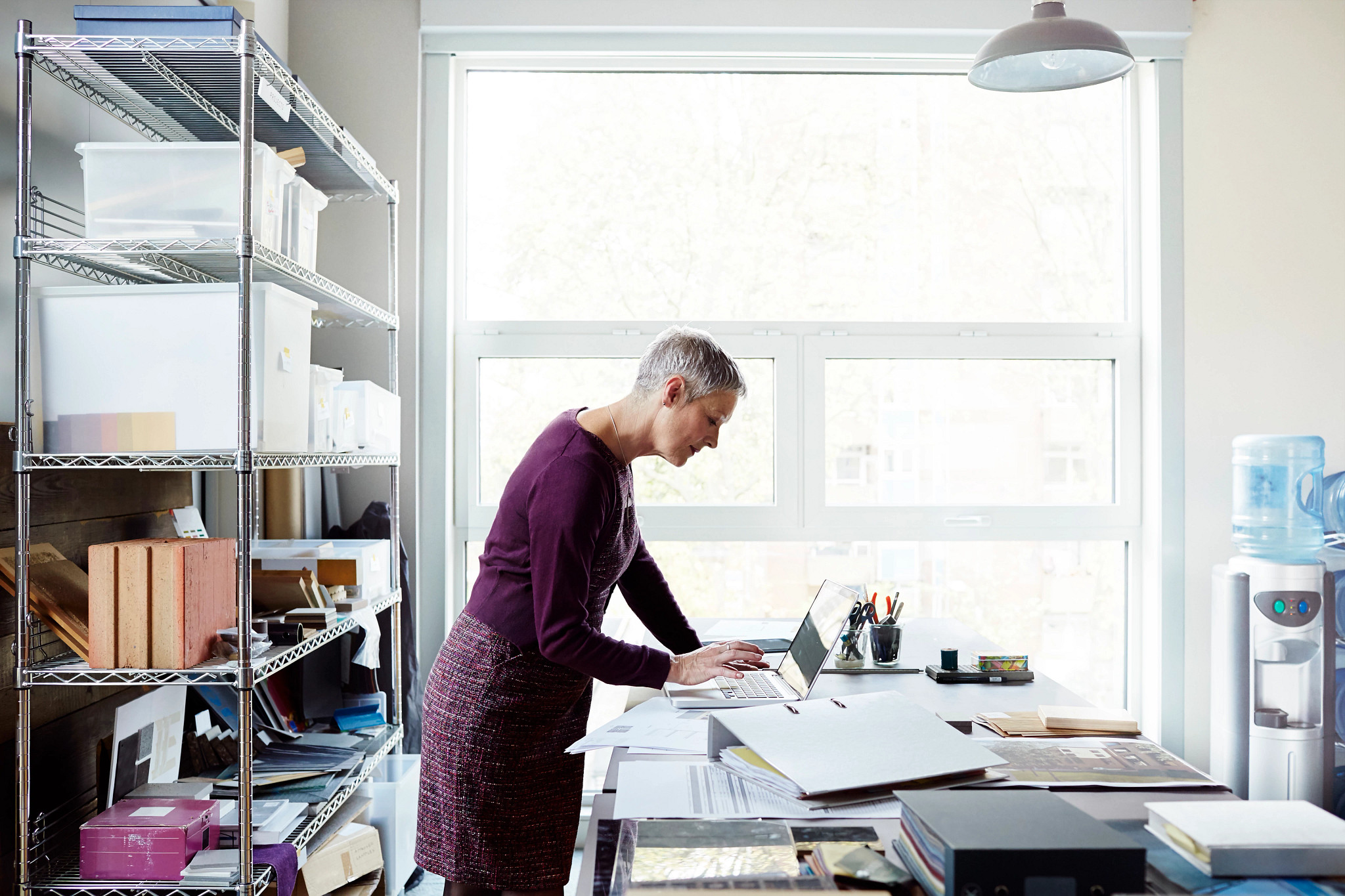 woman with short, grey hair standing in a bright, modern studio or office space, leaning over a large desk to work on a laptop