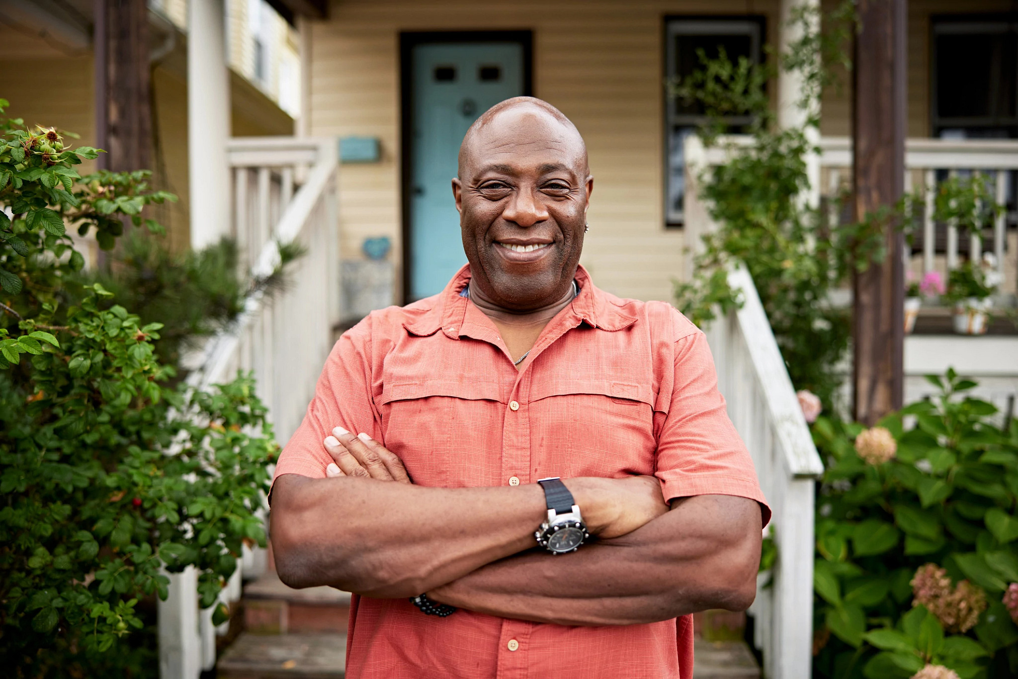 A waist-up view of bald man in late 60s wearing casual open collar shirt, standing with arms crossed and smiling at camera, front porch in background.