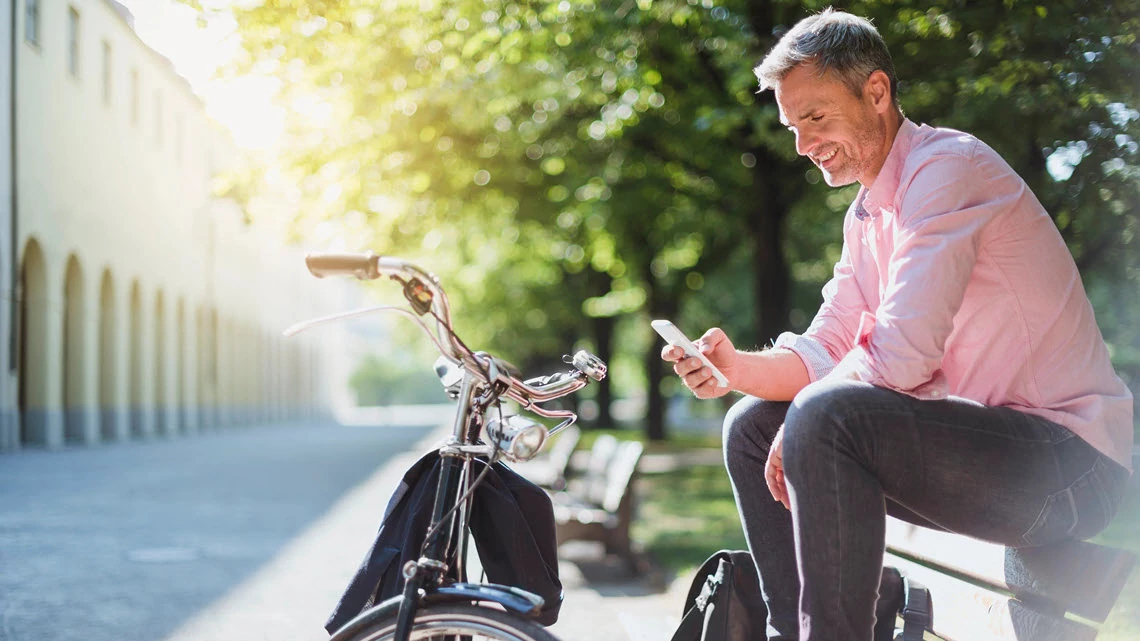 Un  hombre sentado en un banco mirando el teléfono con una bicicleta parada frente a él.