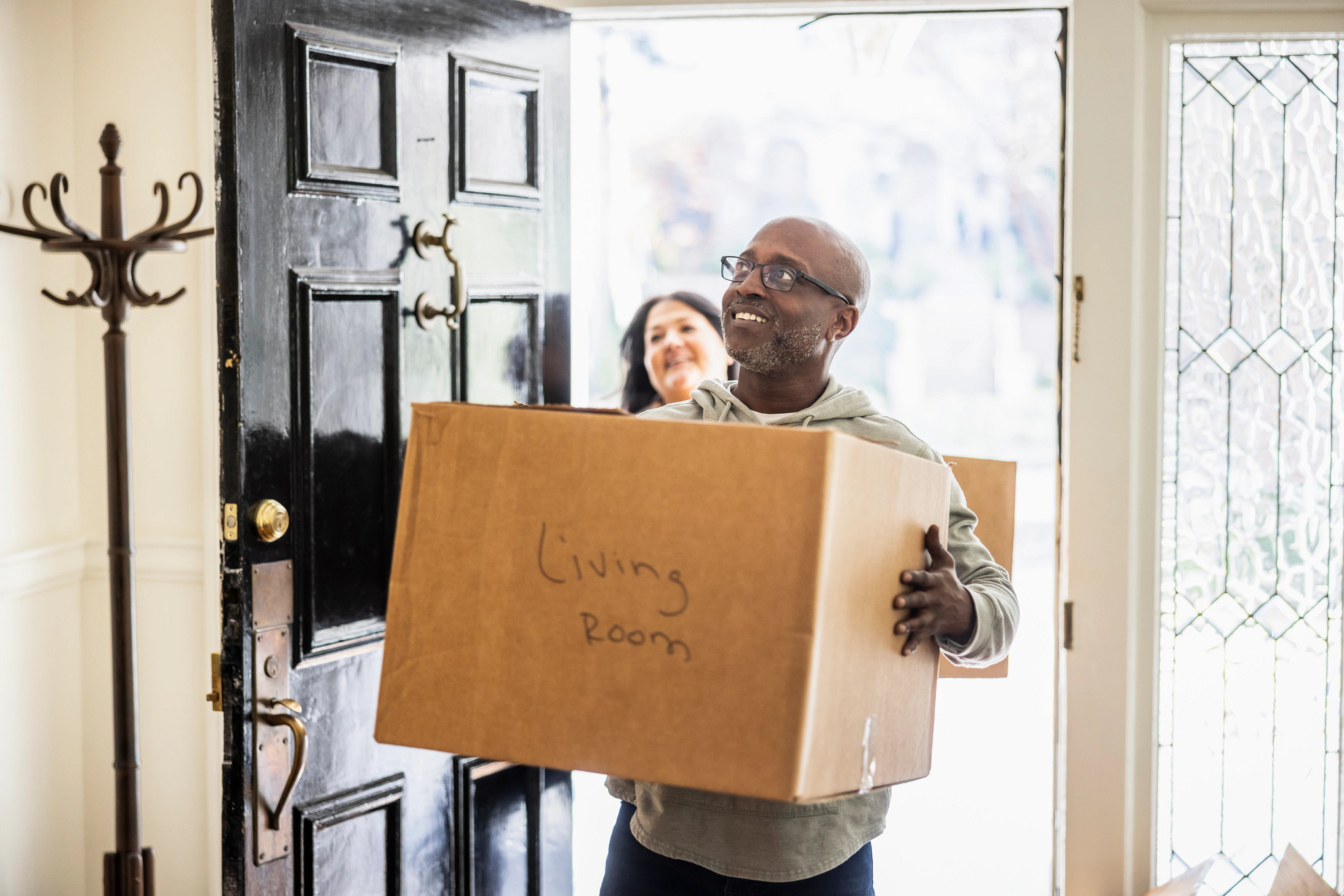 A smiling older adult man carrying a cardboard moving box labeled "Living Room" through the front door of a house as a woman follows behind him.