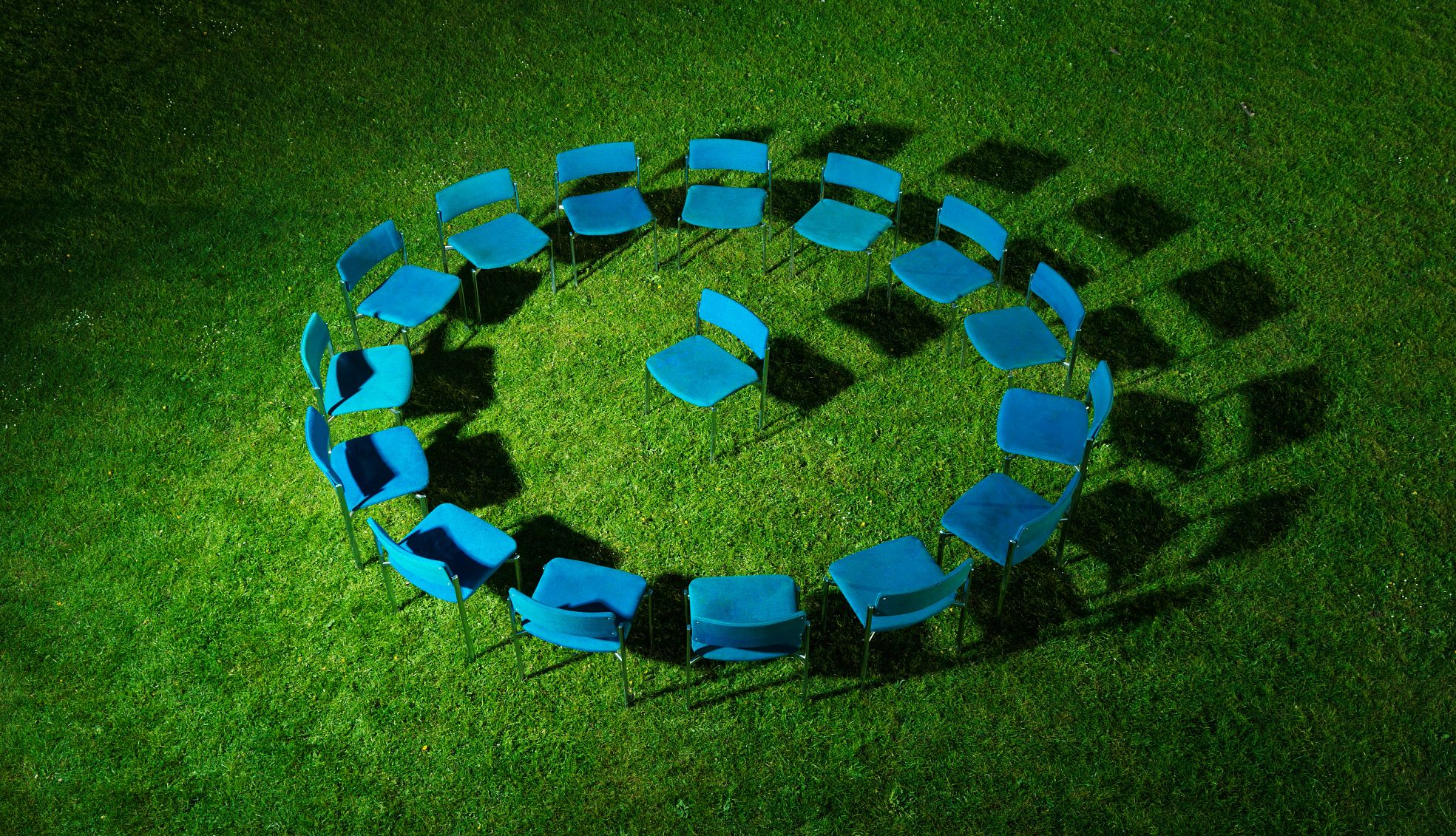 circle of blue chairs on green grass field, one chair in centre, elevated view