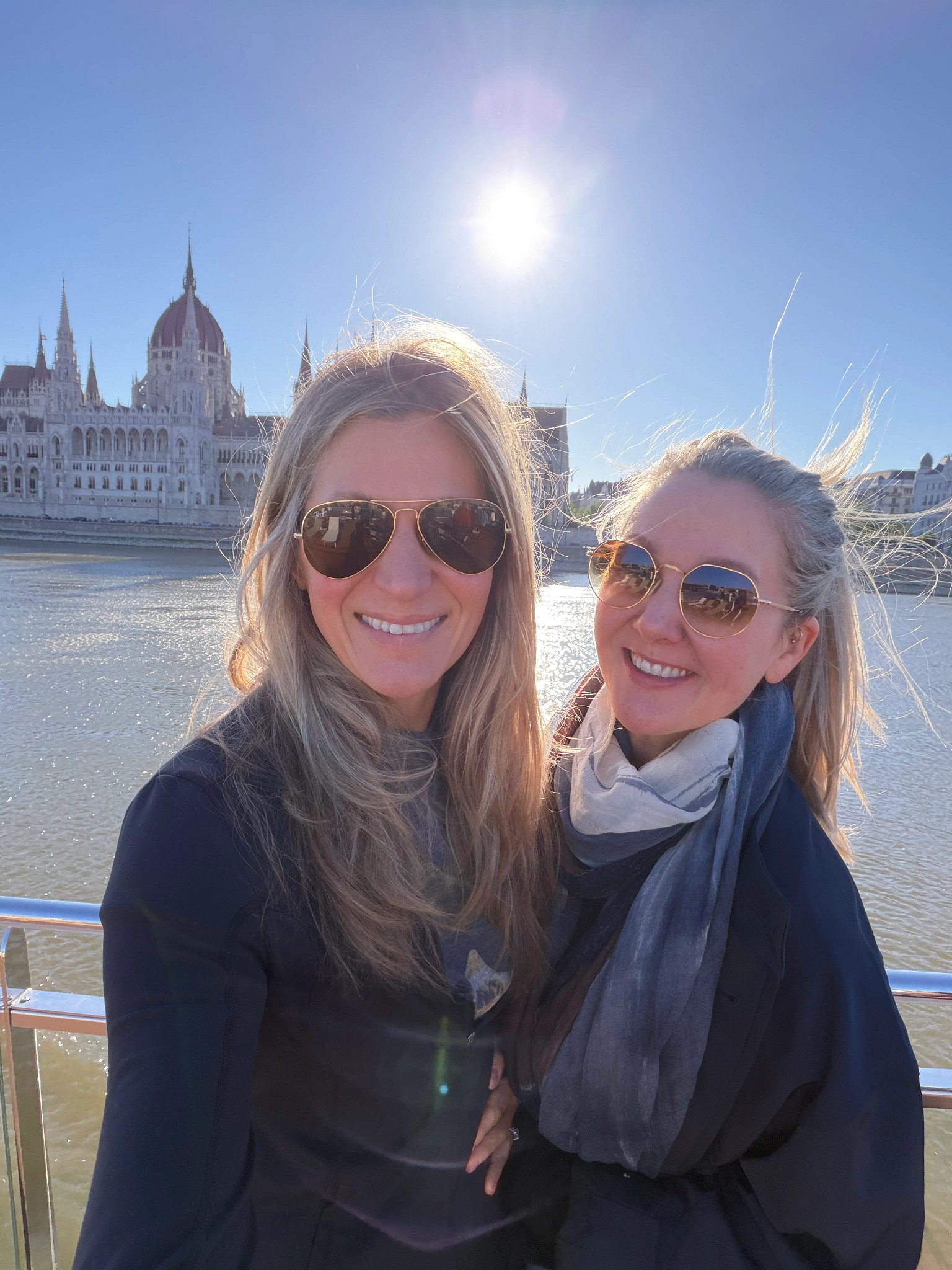 Two adults wearing sunglasses stand on a riverside promenade with water and a historic parliament building in the background on a sunny day