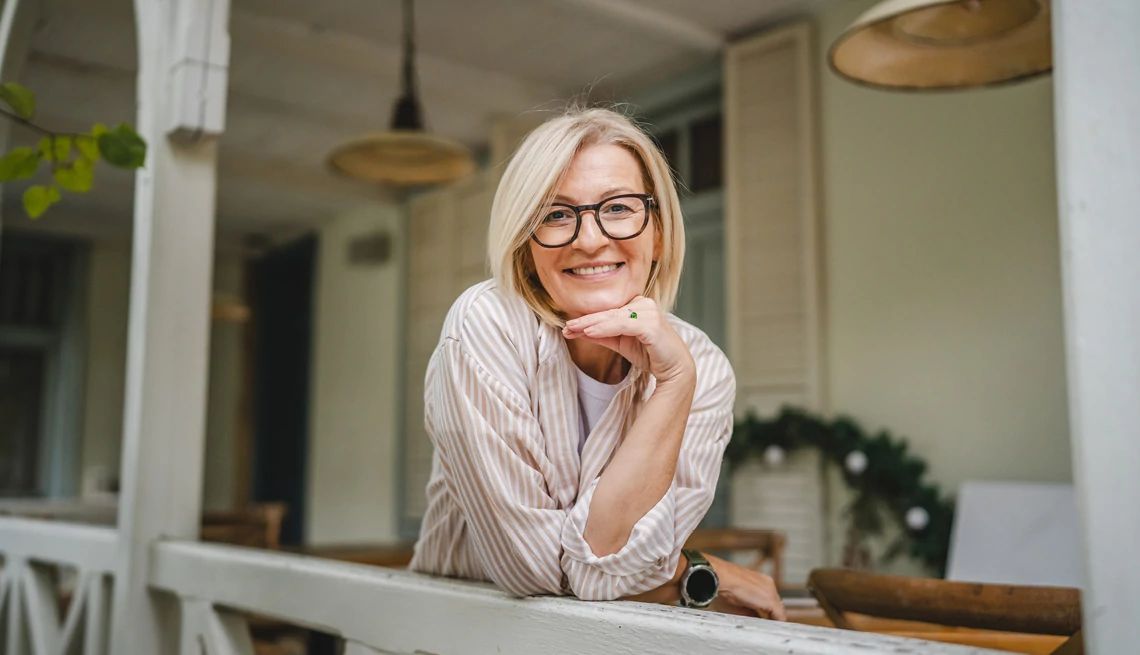 AARP Homeowners Insurance from The Hartford Smiling mature woman leaning on the railing of the porch in front of her home