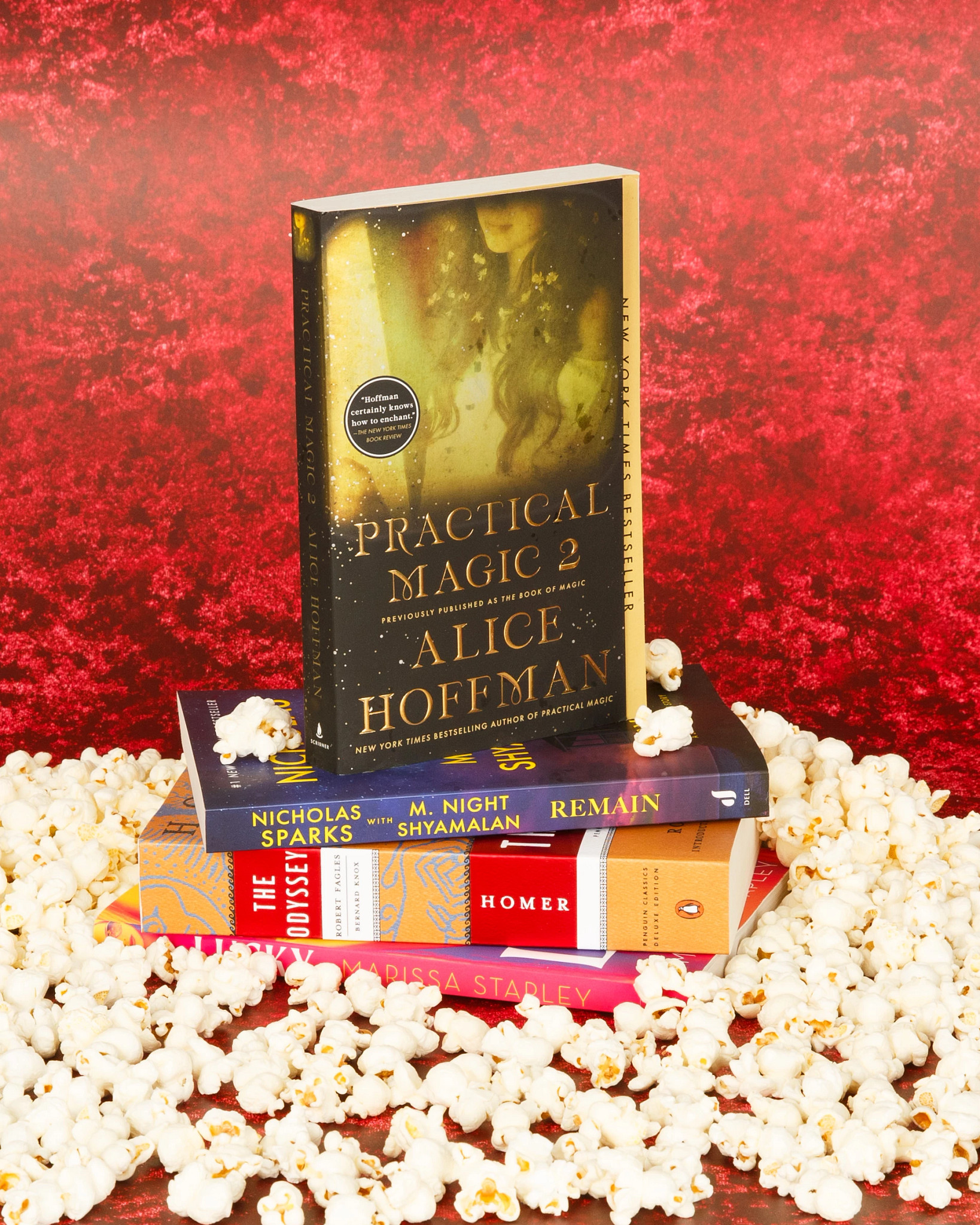A stack of books including “Practical Magic 2” by Alice Hoffman displayed on top of other novels, surrounded by scattered popcorn against a vibrant red background