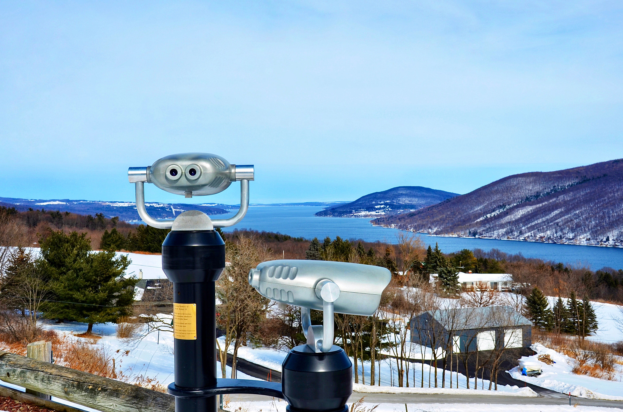 Coin-operated viewing telescopes overlook a scenic lake surrounded by snow-covered hills and lakeside homes.