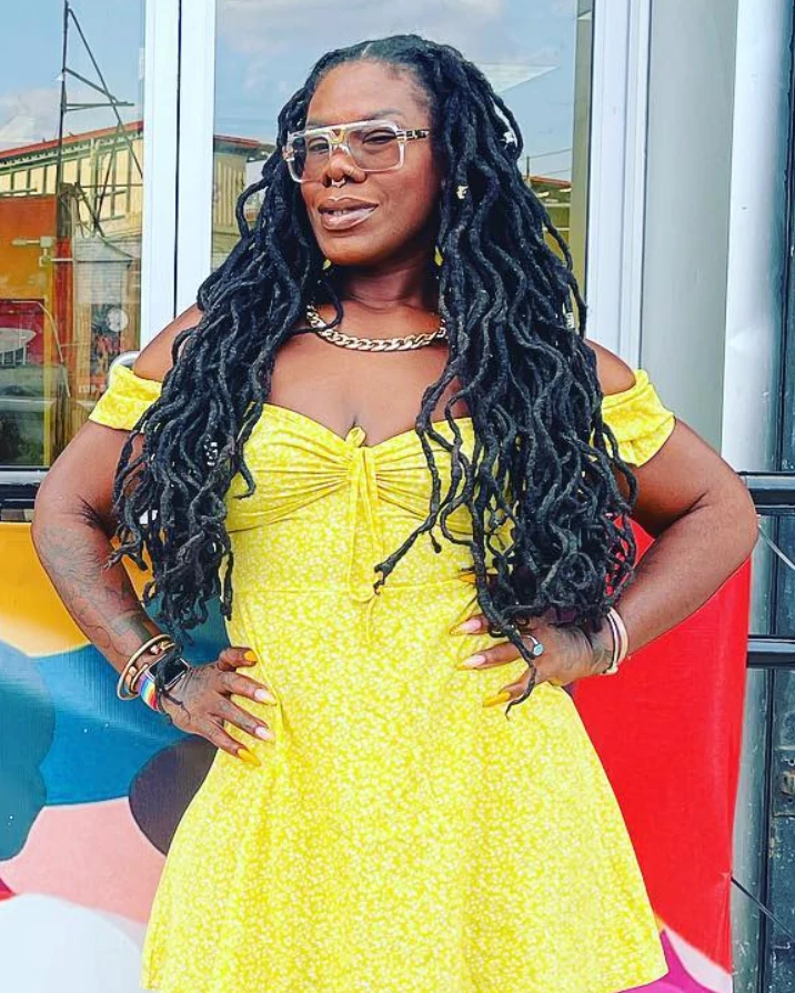 Tanya Fields wearing a bright yellow dress and glasses poses outdoors near colorful artwork