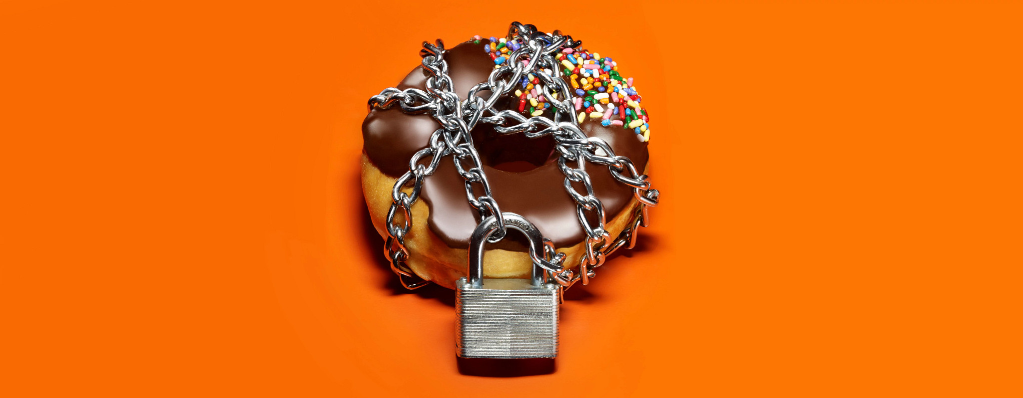 A chocolate frosted donut on an orange background with a chain and lock around it.