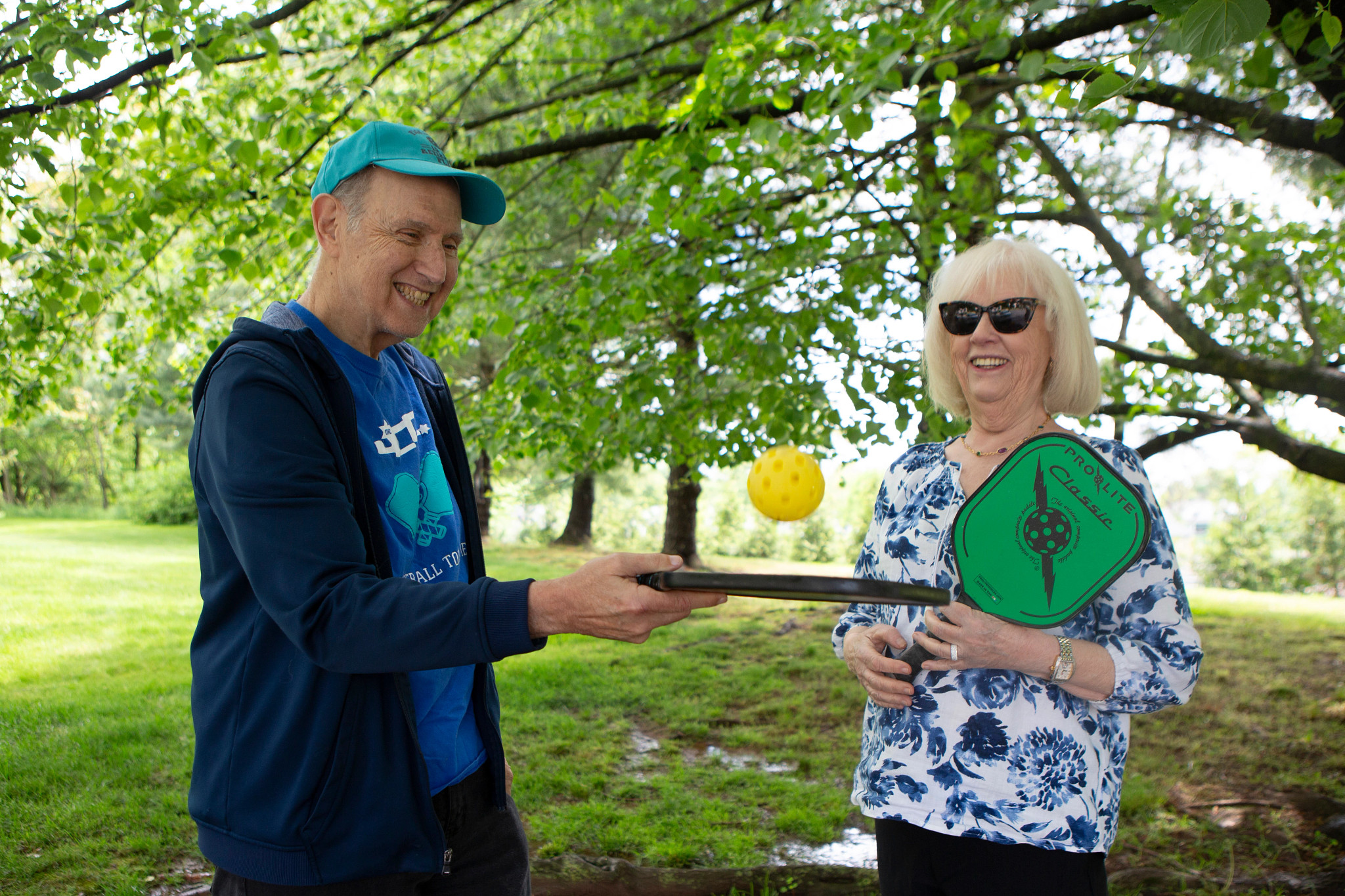 abe rosenstein bounces a yellow ball off his pickleball racquet. next to him, his wife judy smiles while holding a green and black pickleball racquet.
