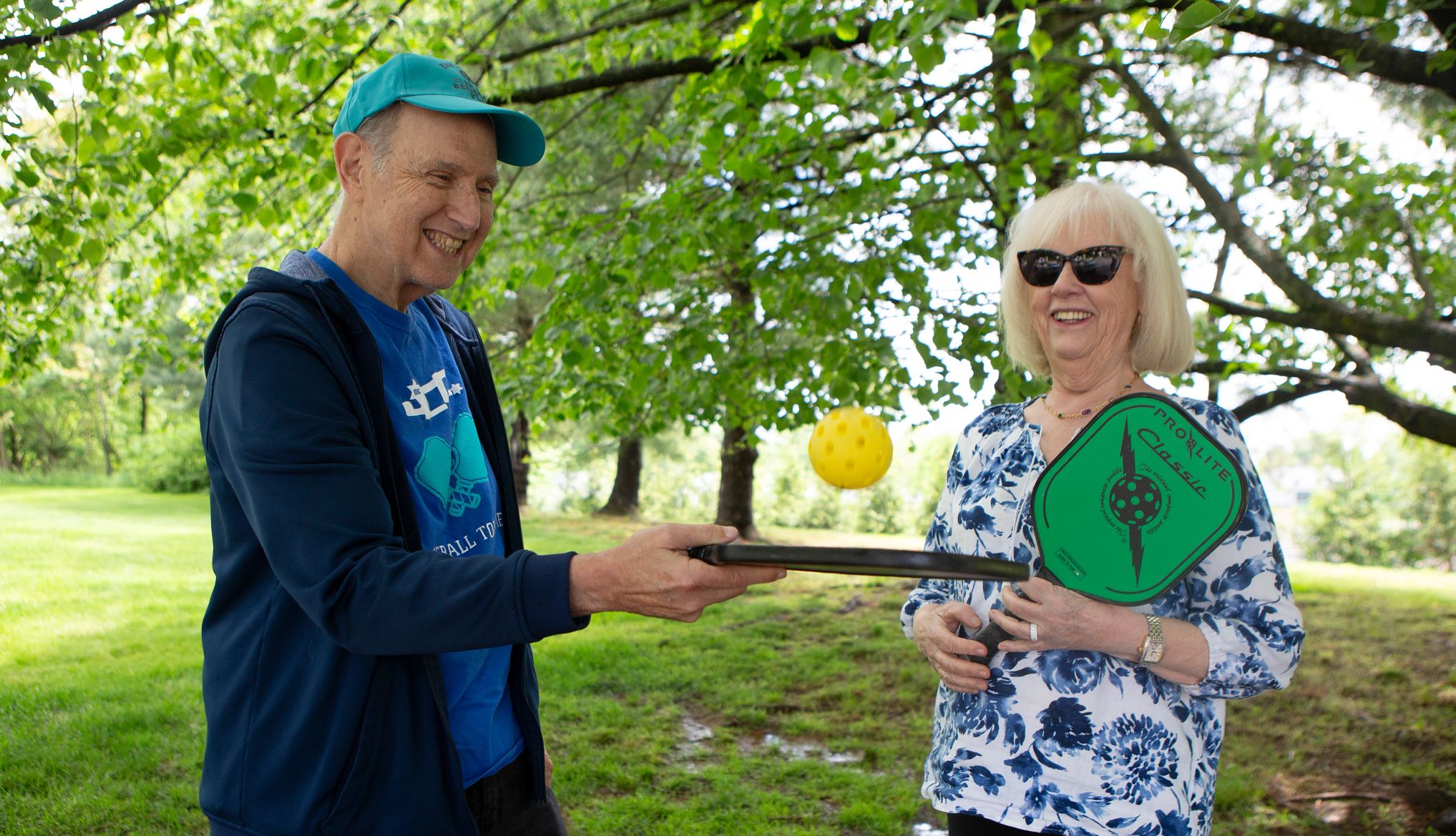 abe rosenstein bounces a yellow ball off his pickleball racquet. next to him, his wife judy smiles while holding a green and black pickleball racquet.