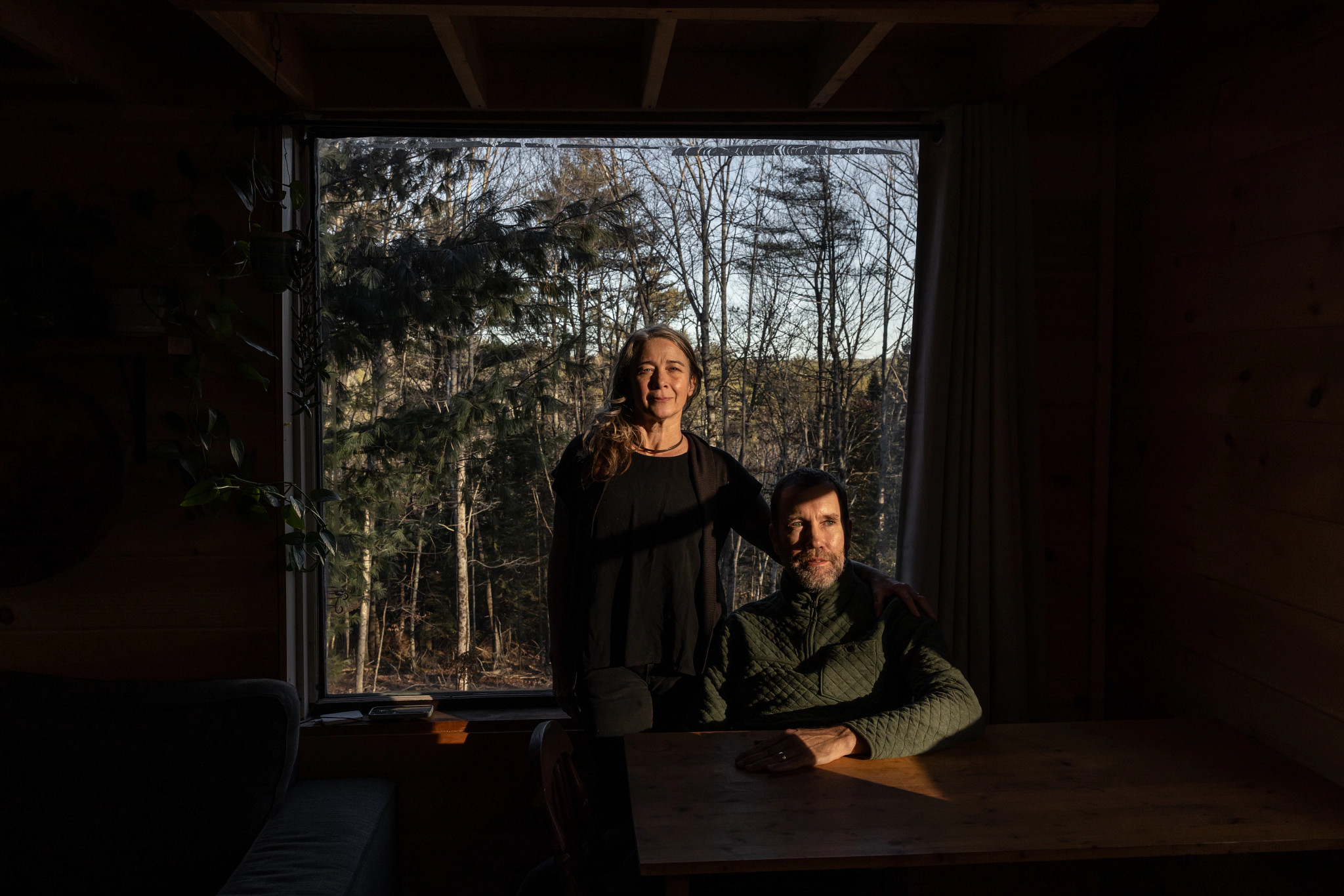 a woman stands behind a man who is sitting at a table. they are in front of a window showing woods and trees outside.