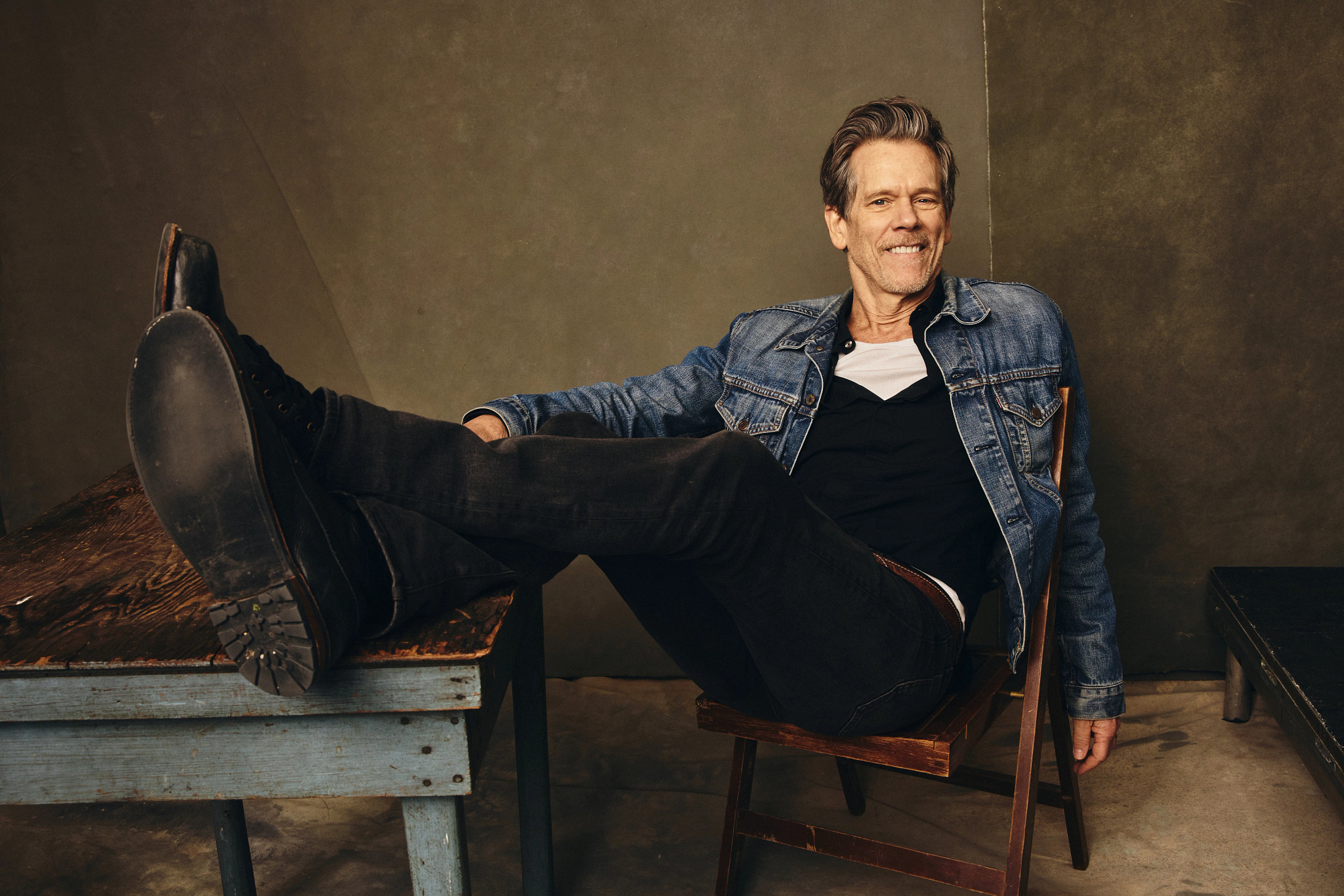 kevin bacon, smiling for a portrait while seated on wooden chair, wearing a casual outfit