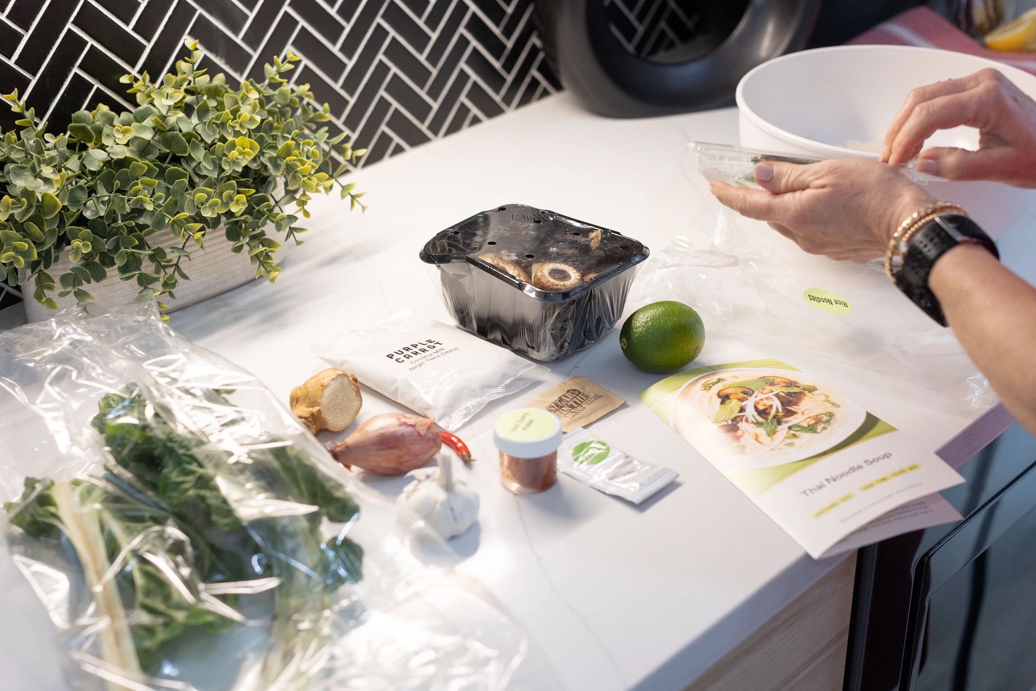 Packaged vegetables and other ingredients on a white counter alongside a plant, a white bowl and the hands of an adult