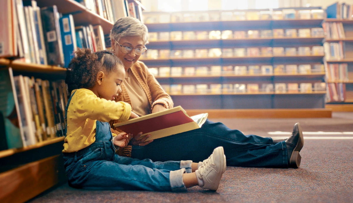 Photograph of an older woman sitting on the floor of a library tutoring a child to read