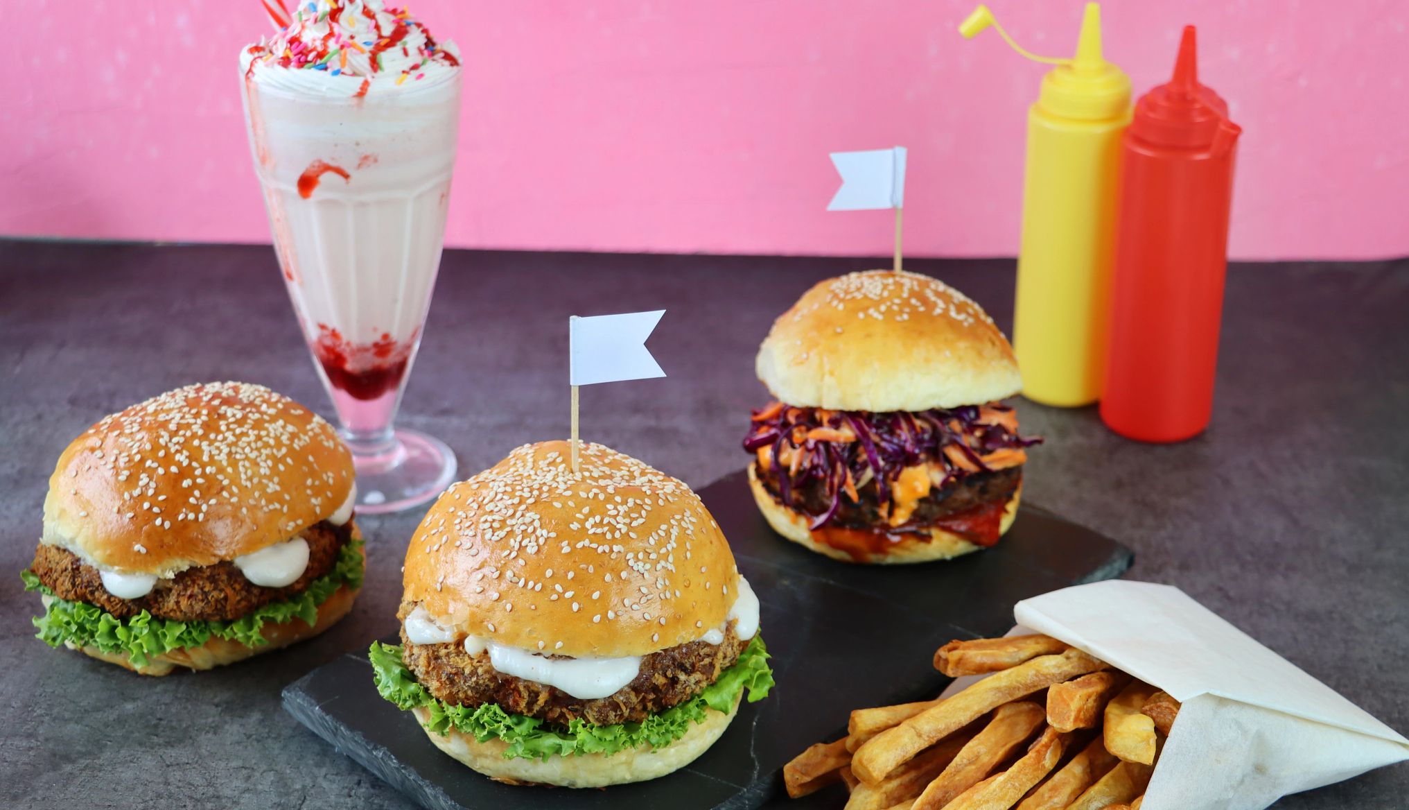 Valentine's Day restaurant deals hamburgers with cheddar cheese, ketchup, red cabbage coleslaw and lettuce, next to french fries, a milkshake and bottles of ketchup and mustard