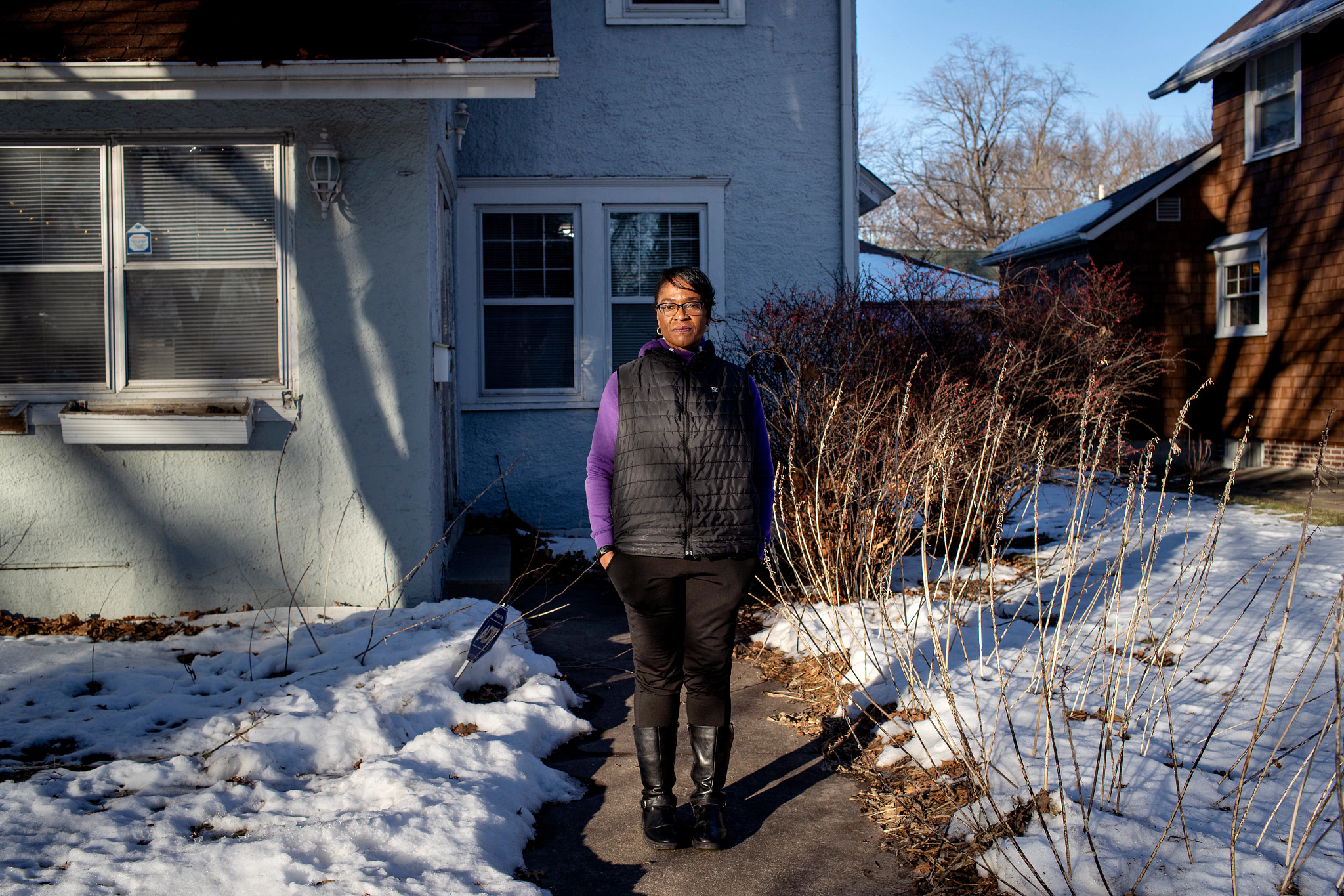 a woman, dressed in black pants, purple shirt and black puffer vest, stands in front of a home in winter