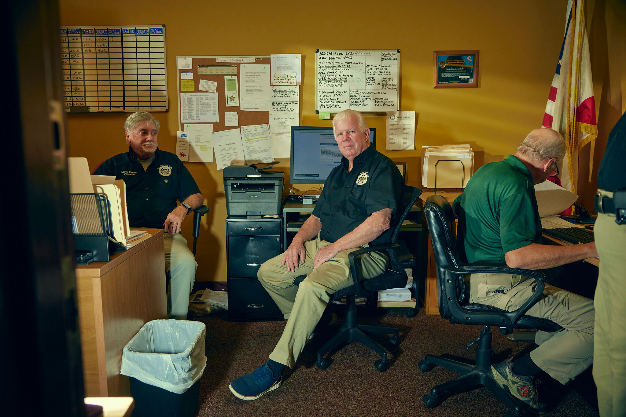 three men sit in an office surrounded by computers and paperwork