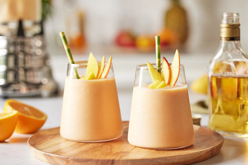 Two creamy peach smoothies with apple slices and green straws on a wooden board
