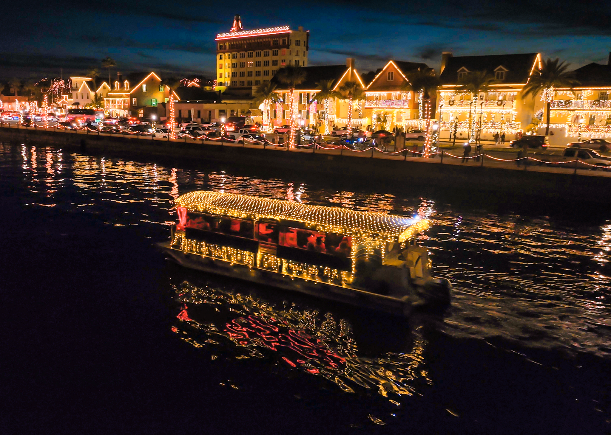a boat with holiday lights travels down a body of water near houses illuminated with lights