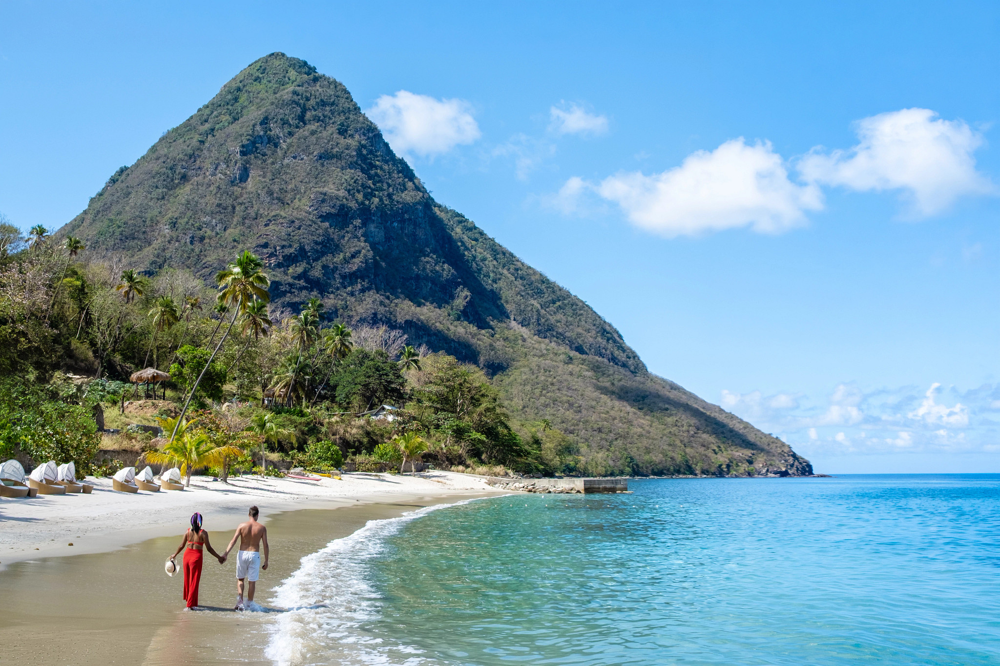 A couple walks along a sunny tropical beach with turquoise water and a towering green mountain in the background