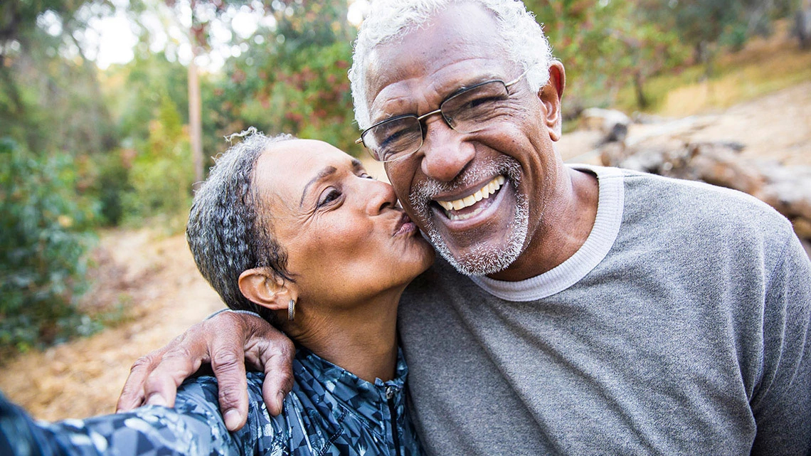 woman and man walking outside, man arm around woman's shoulder, woman kissing man on cheek, he is wearing glasses and smiling