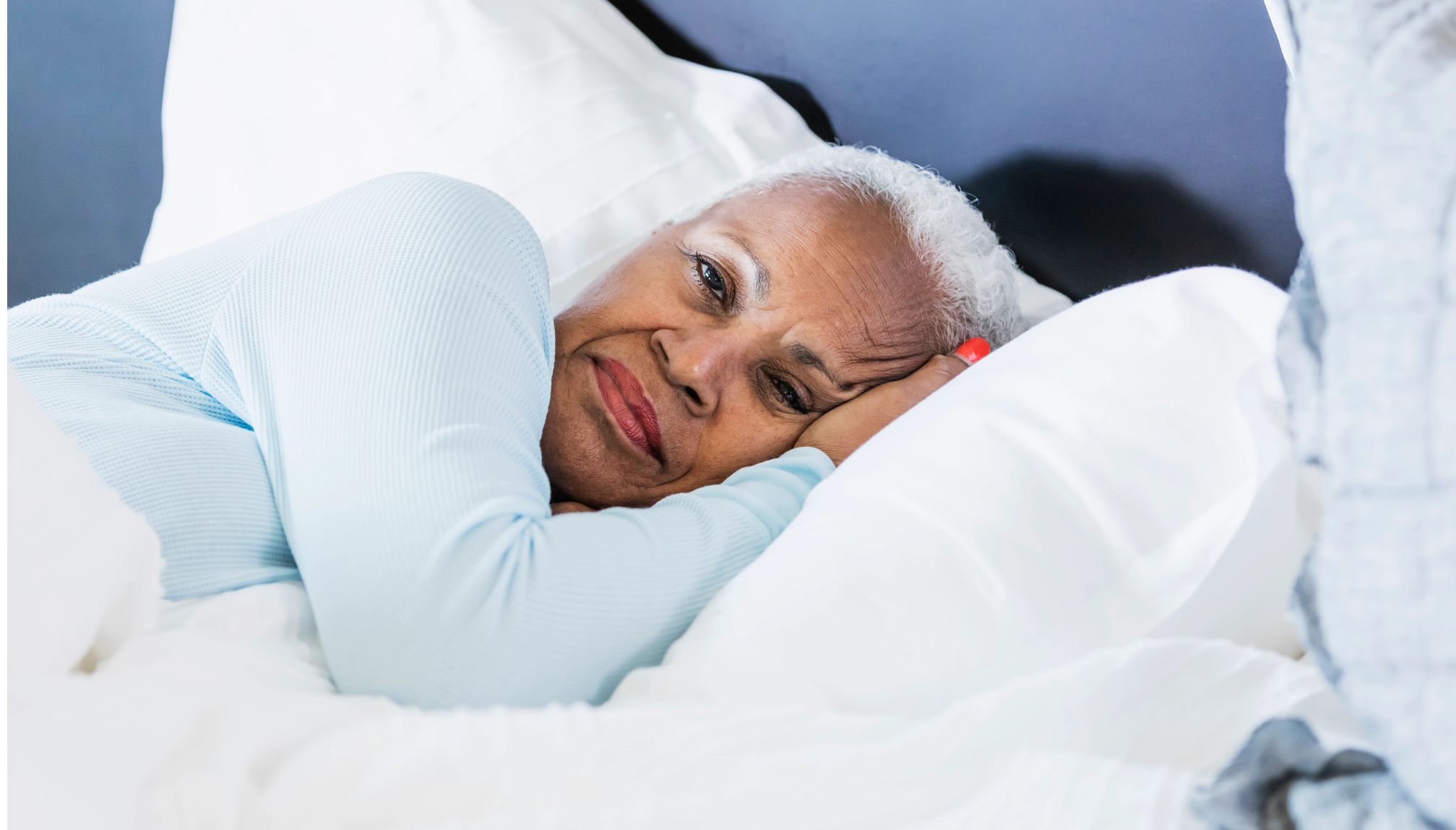A woman in her 60s lying in bed, her side with her eyes open, and a serious expression on her face. It is morning, time to wake up.