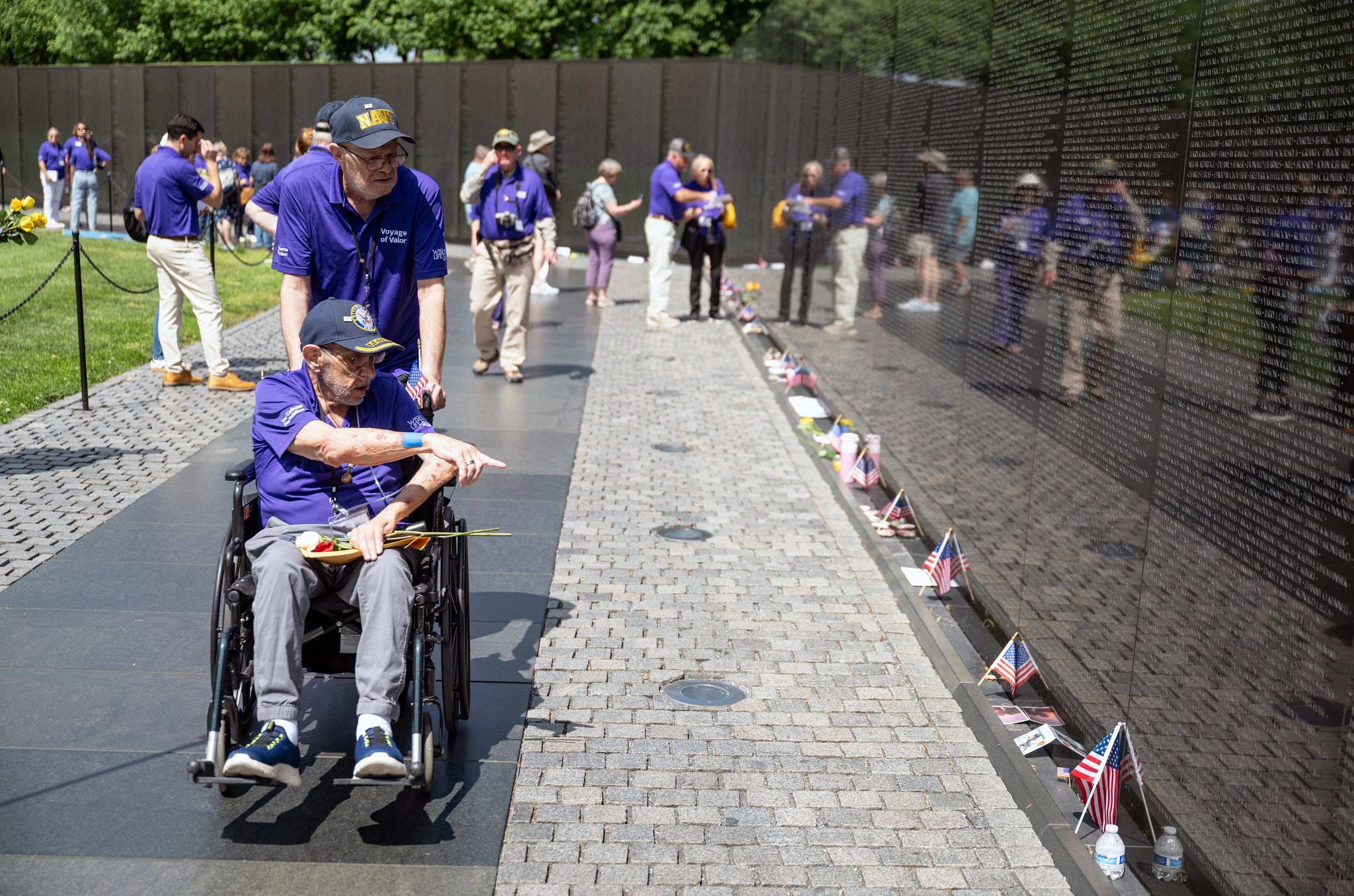LGBTQ Veteran Finds Closure Through Wish of a Lifetime Billy Fiorelli, seated in a wheelchair, pointing at a name on the Vietnam Memorial wall while his husband escorts him