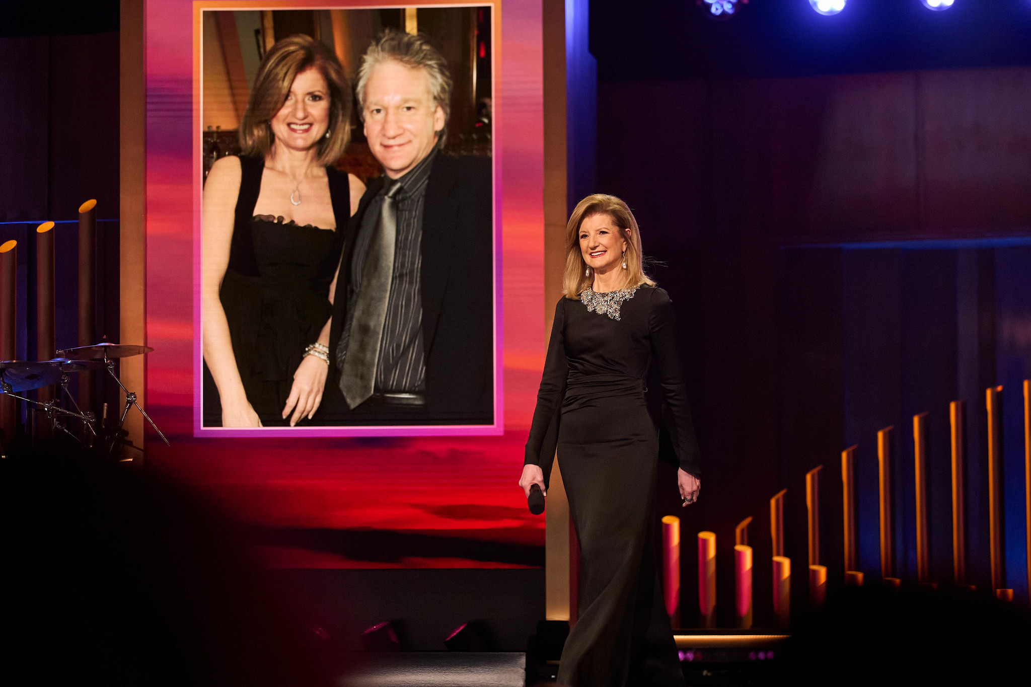 arianna huffington standing on a stage in front of a large projection of a photo of herself and bill maher