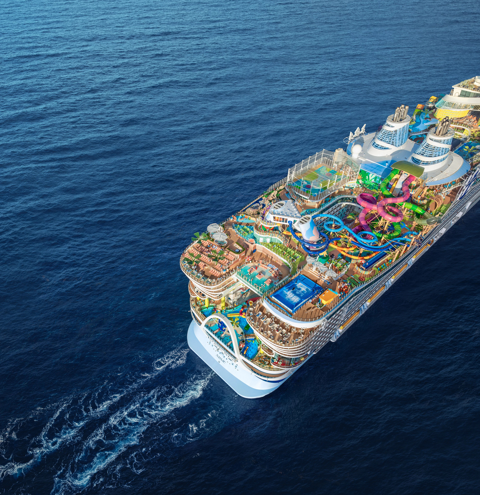 High-angle aerial photograph of a large cruise ship traveling across deep blue ocean waters. The vessel features expansive pool decks, colorful waterslides, sports courts, and outdoor recreation areas