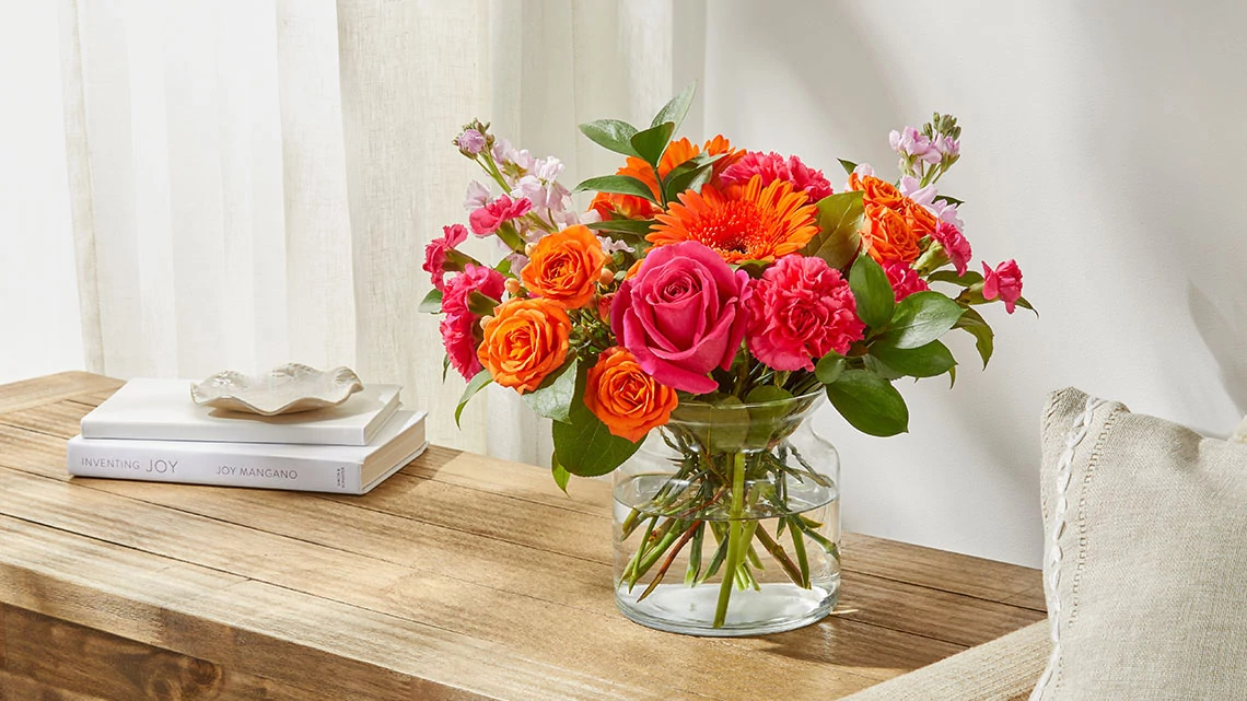 Flowers by FTD orange gerber daisies and roses with dark pink roses and carnations and other white, light purple flowers in a vase sitting on a wood table next to a linen colored chair