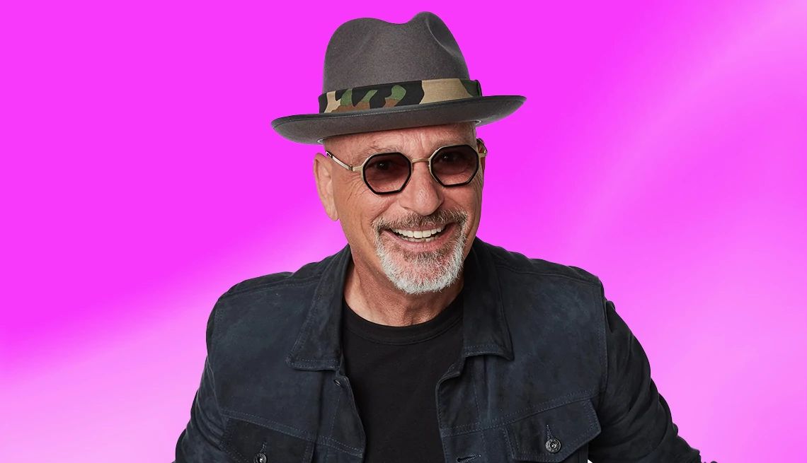 A smiling Howie Mandel wearing a hat and sunglasses against a purple background