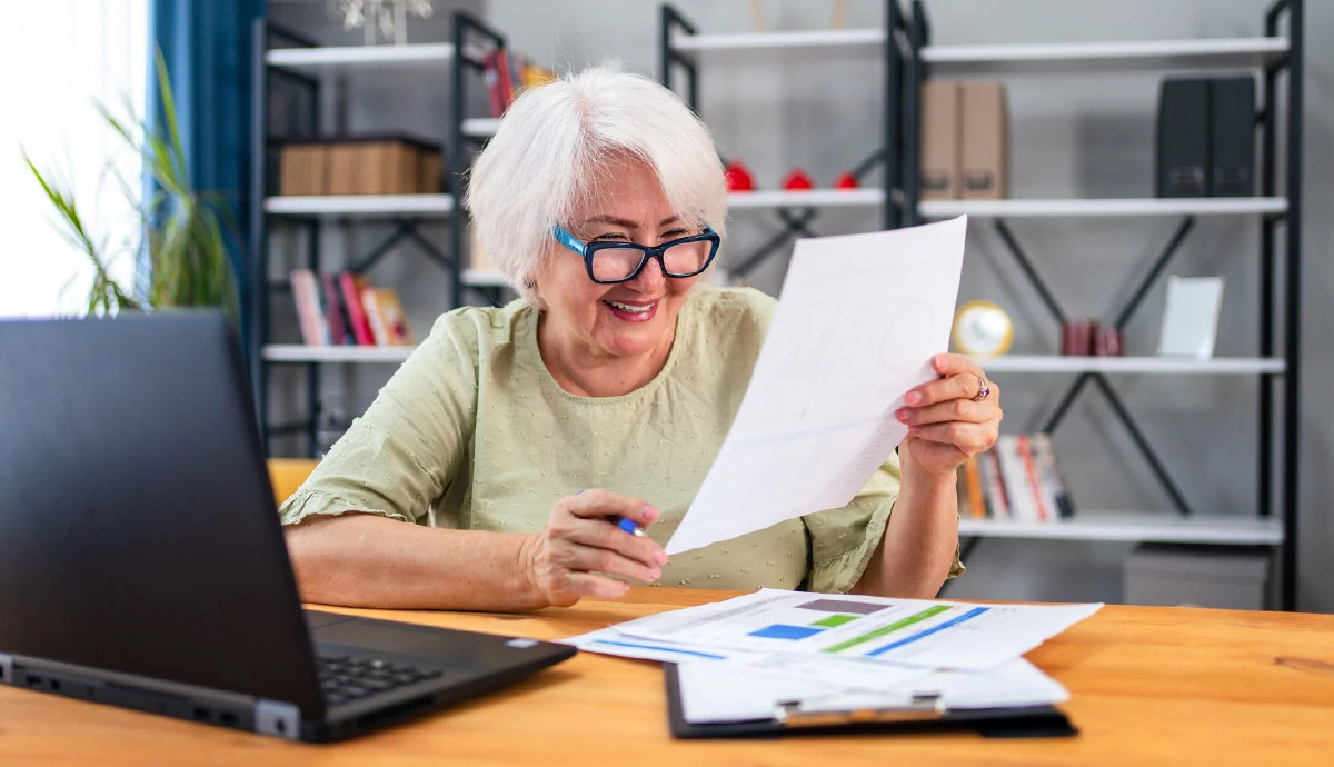A mature woman sitting at a desk in her home paying bills on her laptop.