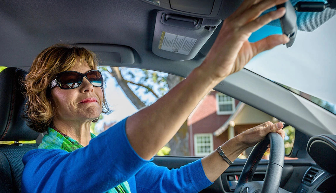 Woman adjusting mirror in car, she is wearing sunglasses and long sleeve blue sweater