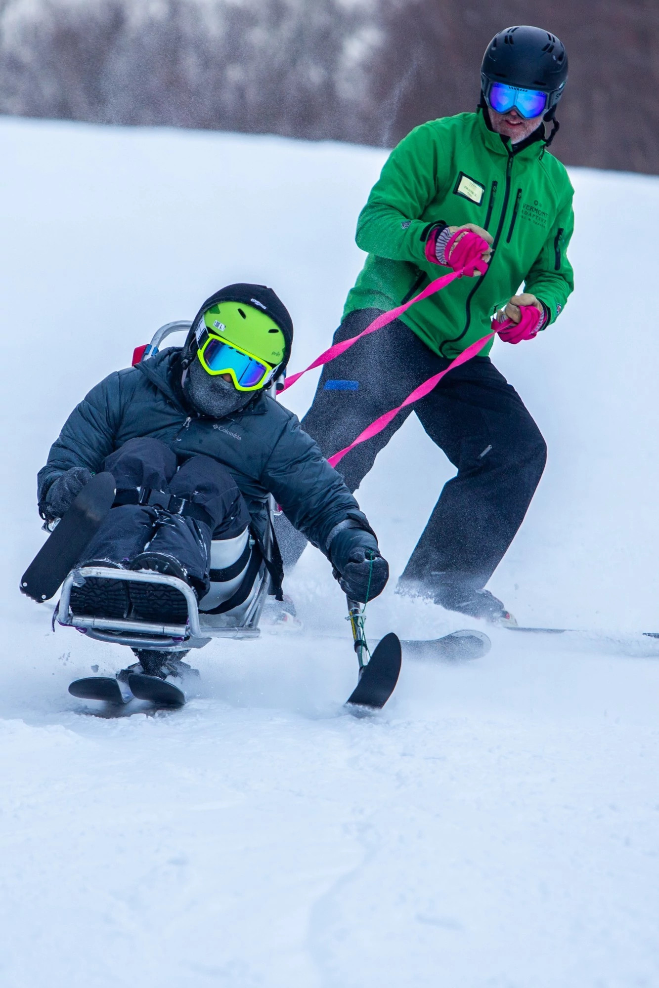 Adaptive skier in a sit-ski is guided down a snowy slope by an instructor holding bright pink straps