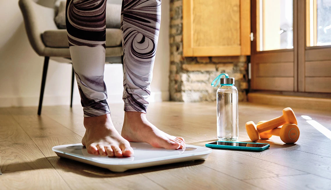 Woman's legs and feet showing standing on a scale with orange hand weights next to the scale with a water bottle and cellphone in a turquoise case, wood floor and window/door in background