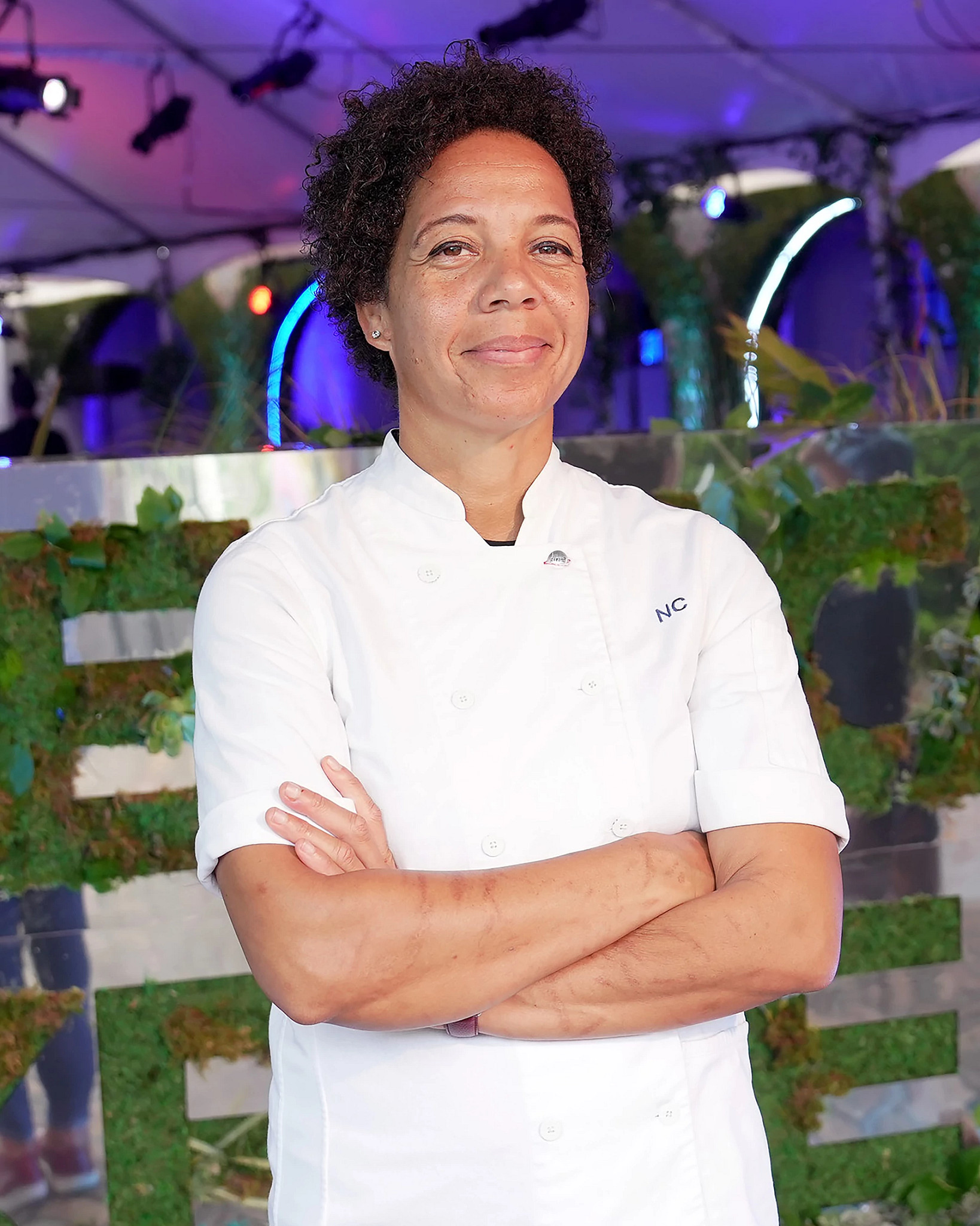 Chef Nina Compton standing with arms crossed in a modern event space with plants and colorful lighting