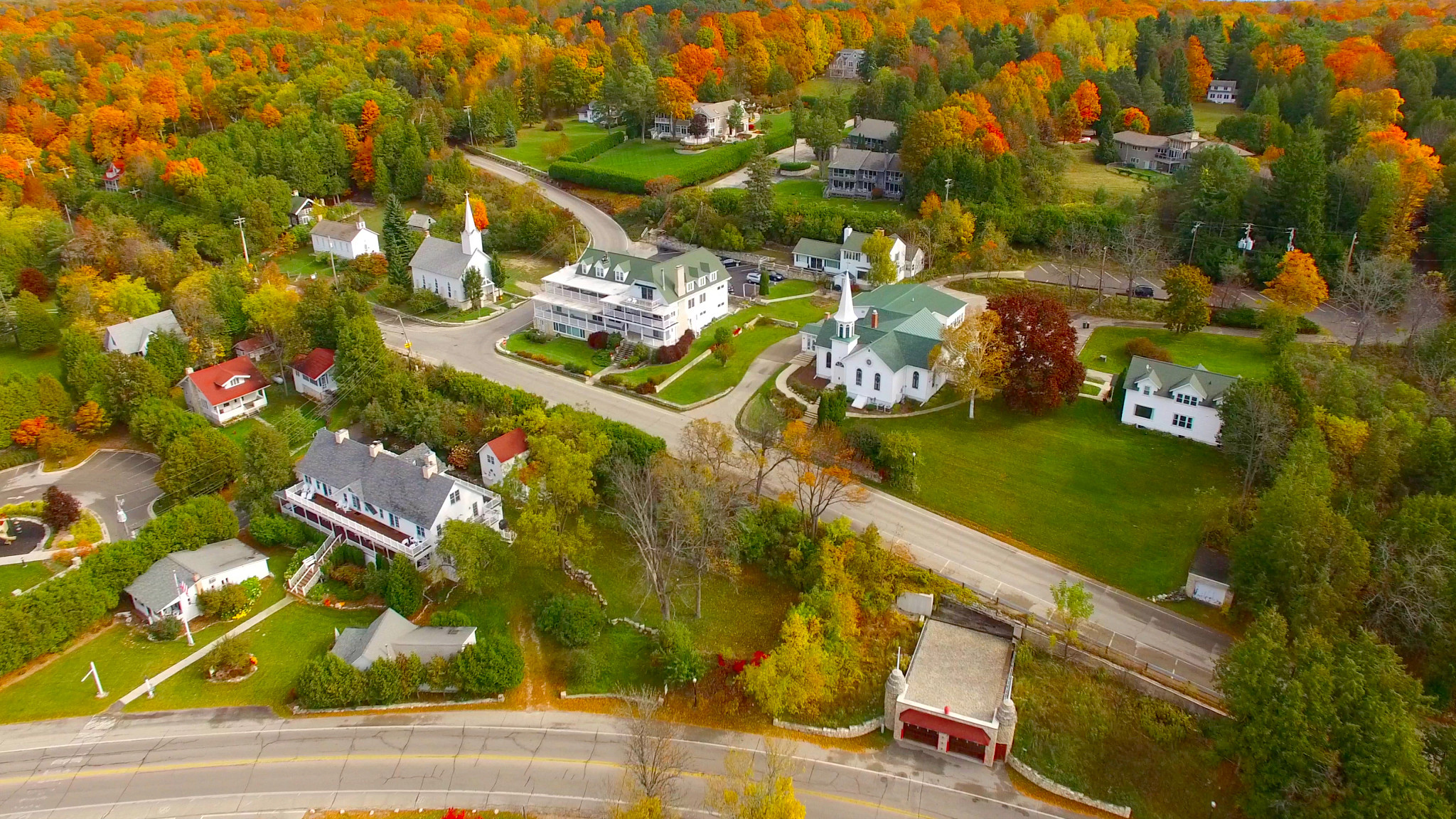 Aerial photograph of a town surrounded by vibrant autumn foliage in shades of orange, yellow, green, and red. Churches, homes, winding roads, and community buildings are nestled within a forested landscape