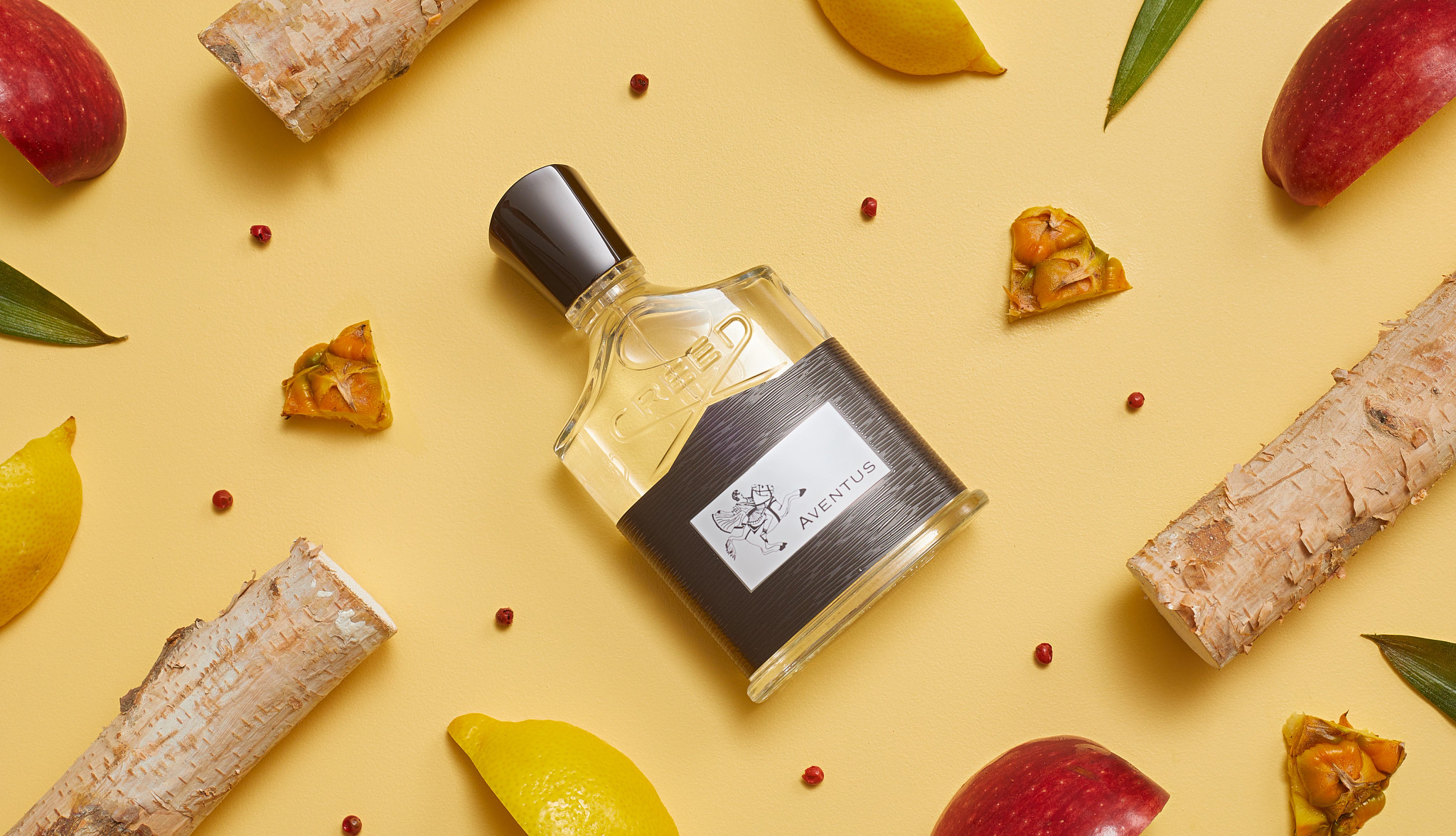 Bottle of Creed Aventus surrounded by lemons, pink pepper, apple slices, bits of pineapple rind, and pieces of birch wood