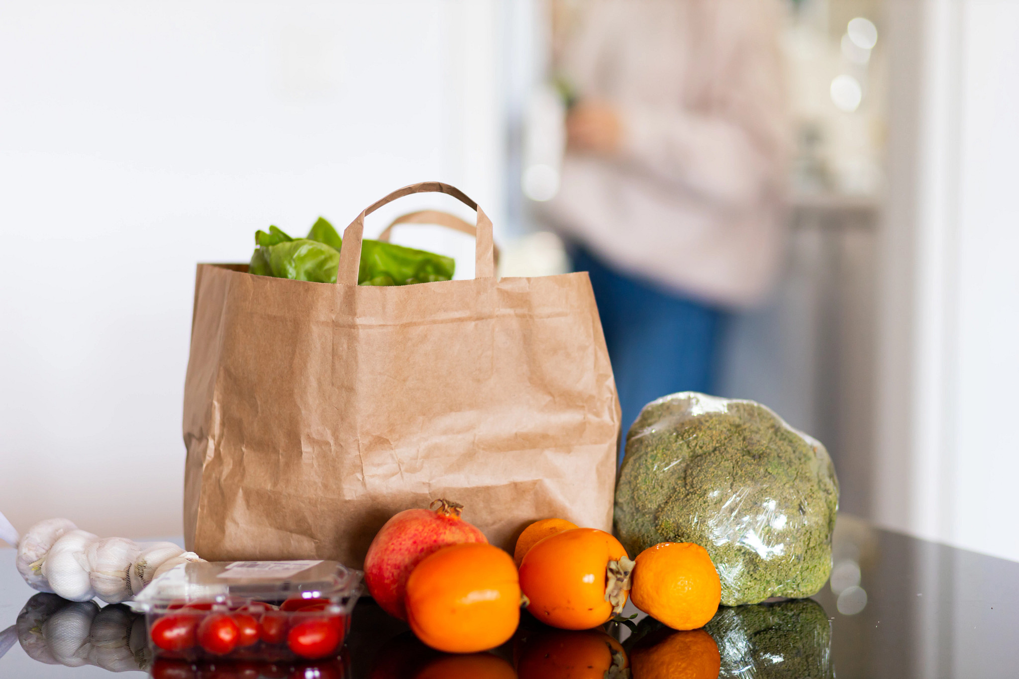 A kitchen counter is pictured with a paper grocery bag. Beside the bag are an array of items, including garlic gloves, tomatoes, cherry tomatoes, a pomegranate, an orange and a head of broccoli.
