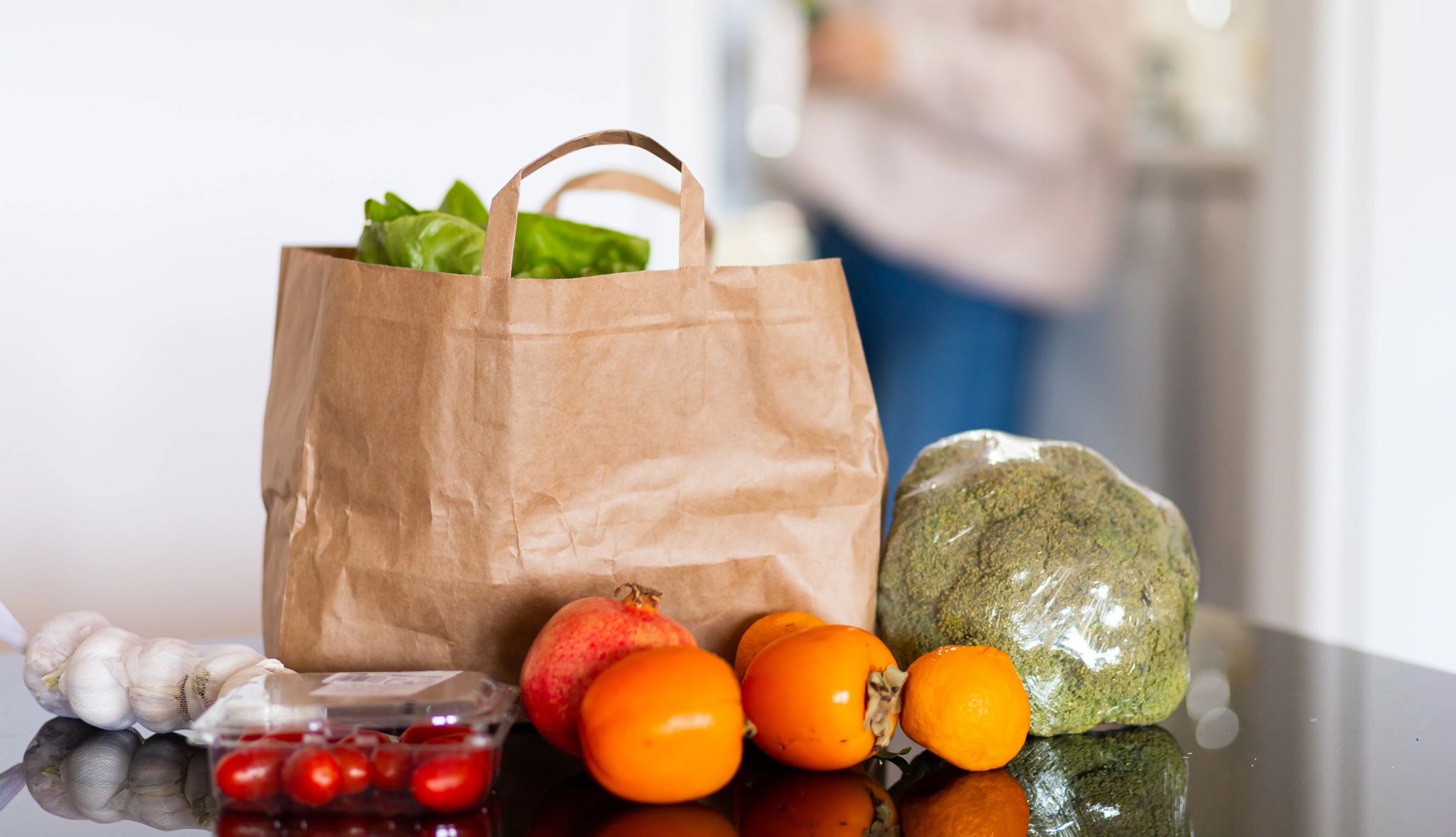 Completing the Plan at Home A kitchen counter is pictured with a paper grocery bag. Beside the bag are an array of items, including garlic gloves, tomatoes, cherry tomatoes, a pomegranate, an orange and a head of broccoli.
