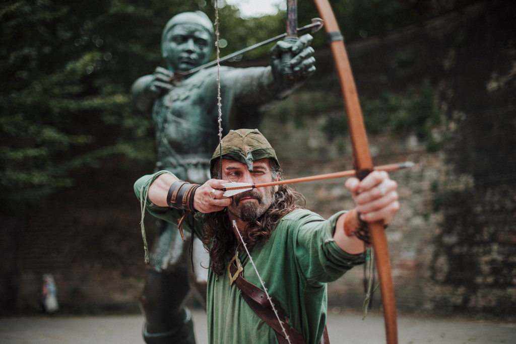 a robin hood performer holds a bow and arrow in front of a robin hood statue
