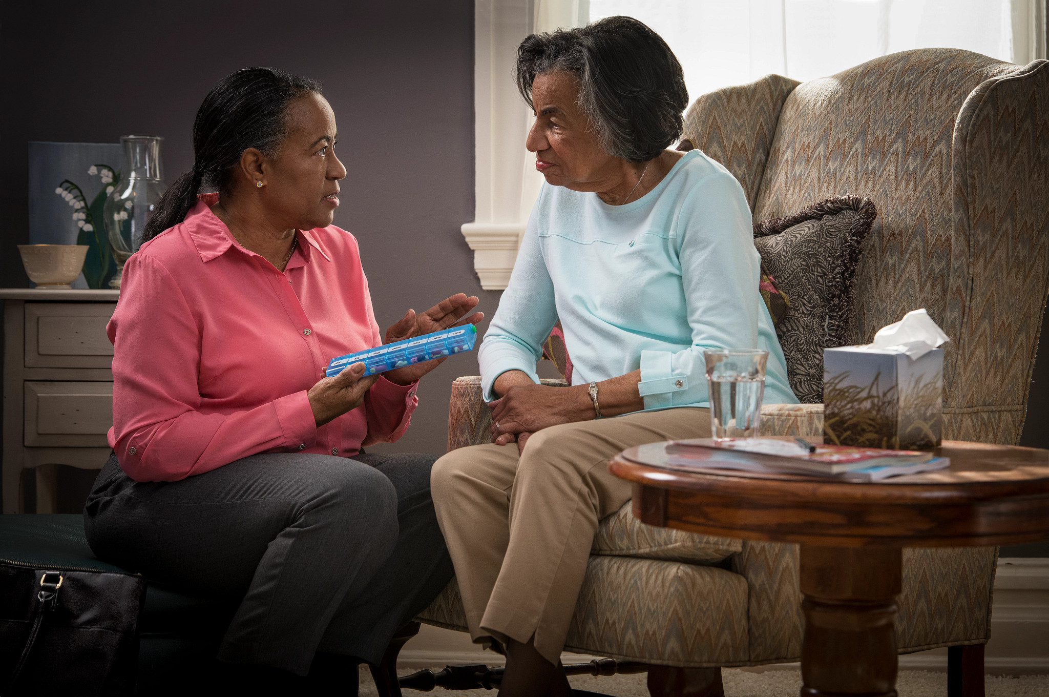 An African American daughter dressed for work kneels in front of her seated African American mother with a pill case.
