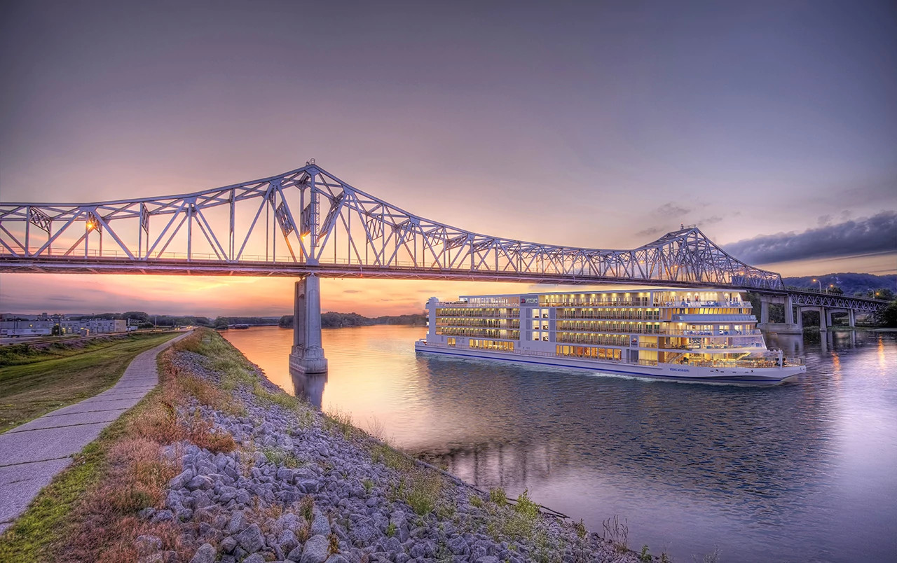 Multi-deck river cruise ship sailing beneath an illuminated steel bridge at sunset
