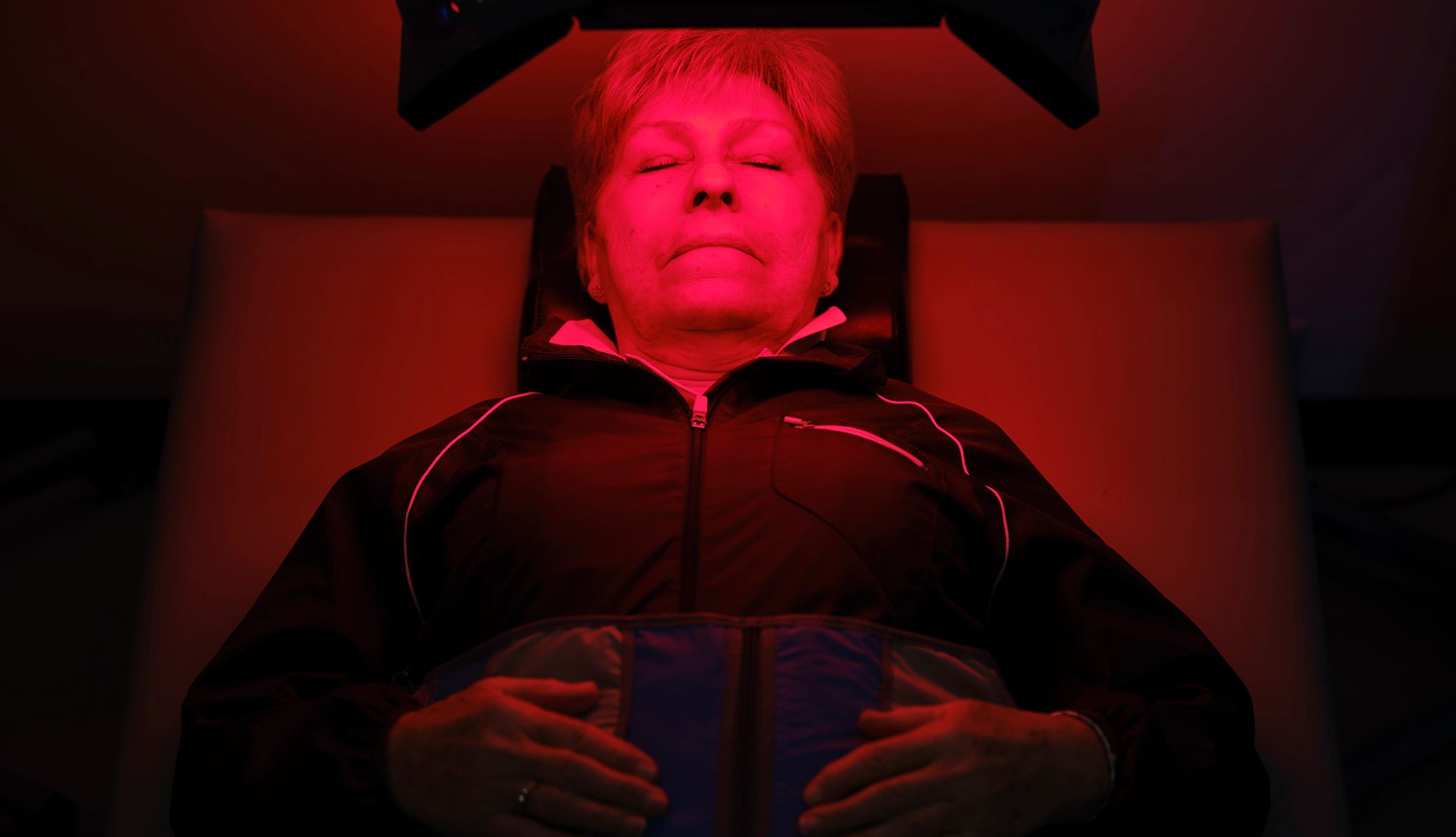 Woman On an LED Bed Receiving Red Light Therapy Una señora mayor reposa bajo una luz roja.