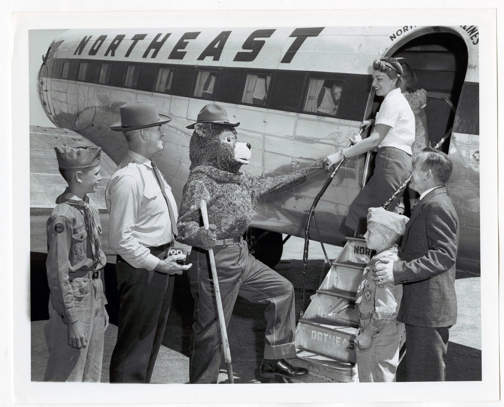 Smokey Bear is shown in a black and white photo greeting travelers as they exit and airplane