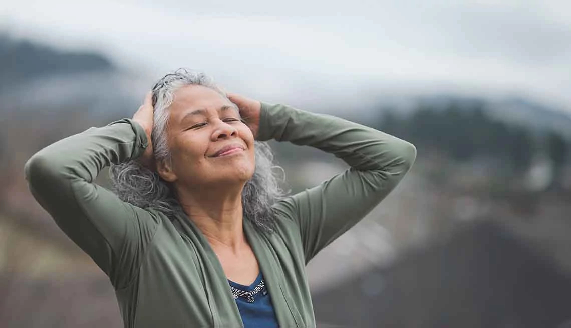 Calm Sleep, Meditation & Relaxation App Woman with gray, long, curly hair, outside with mountains in the background, standing, holding arms up while grabbing top of head, with blissful closed-lips smile, eye closed, head tilted up