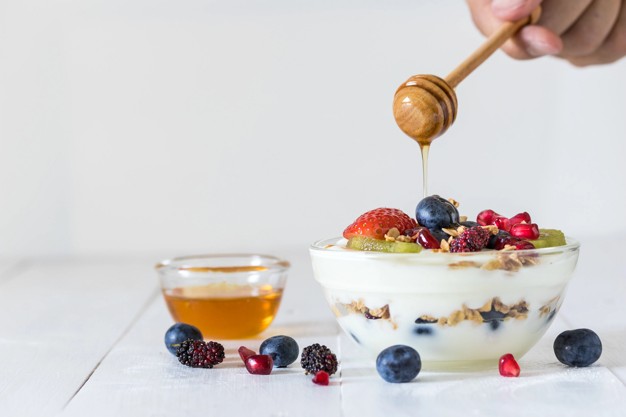 Honey drizzled from a wooden dipper into a glass bowl of yogurt topped with fresh berries, kiwi slices, pomegranate seeds, and granola.